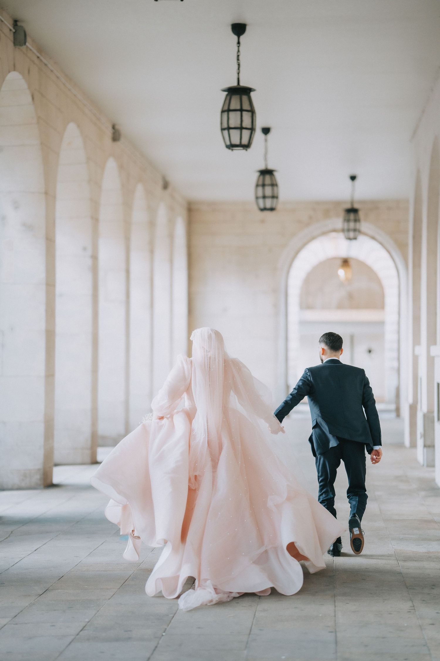 Couple walking through an arched colonnade with flowing blush wedding dress.