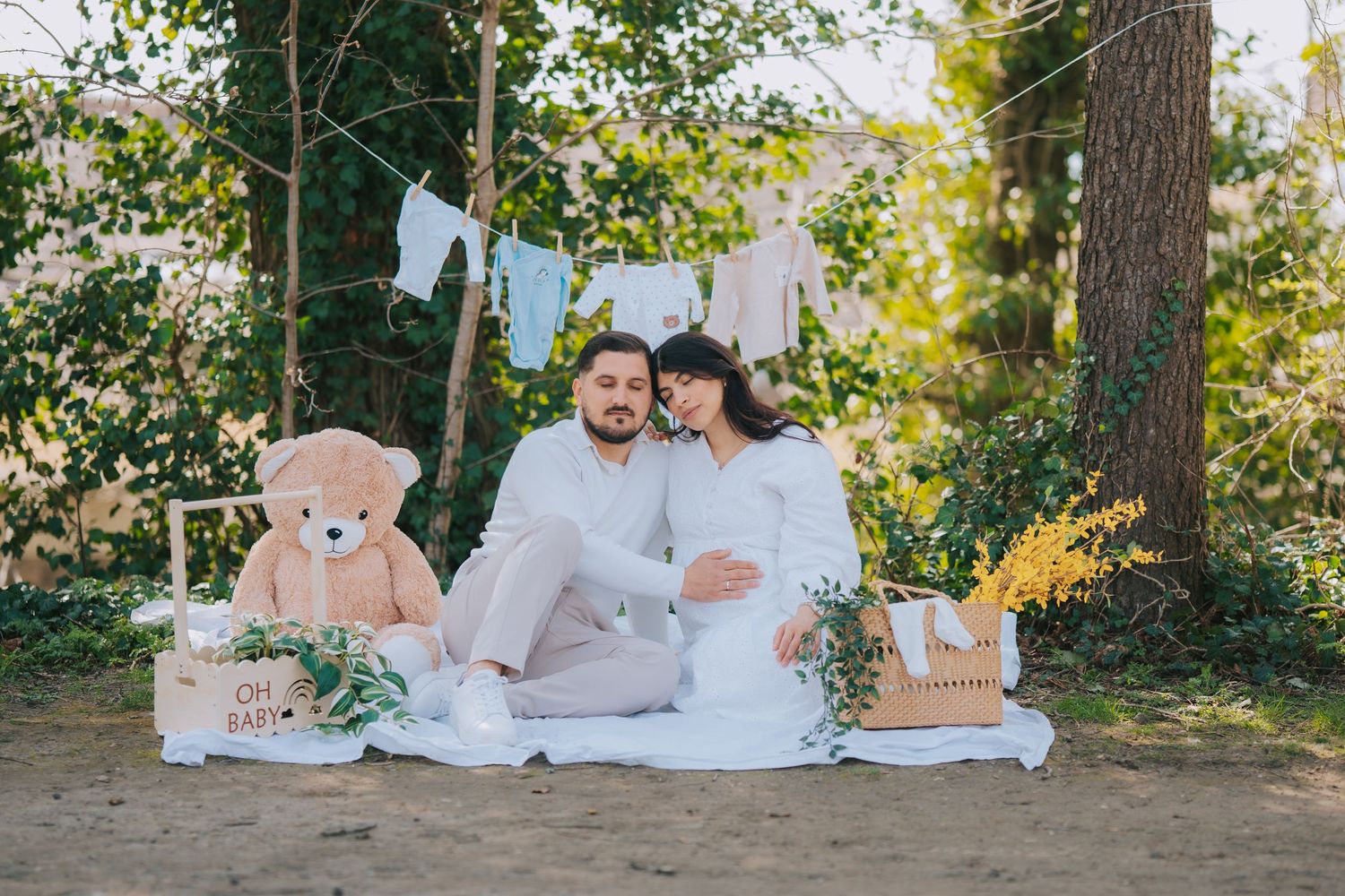 Couple in white outfits sitting on blanket with teddy bear and baby clothes display.