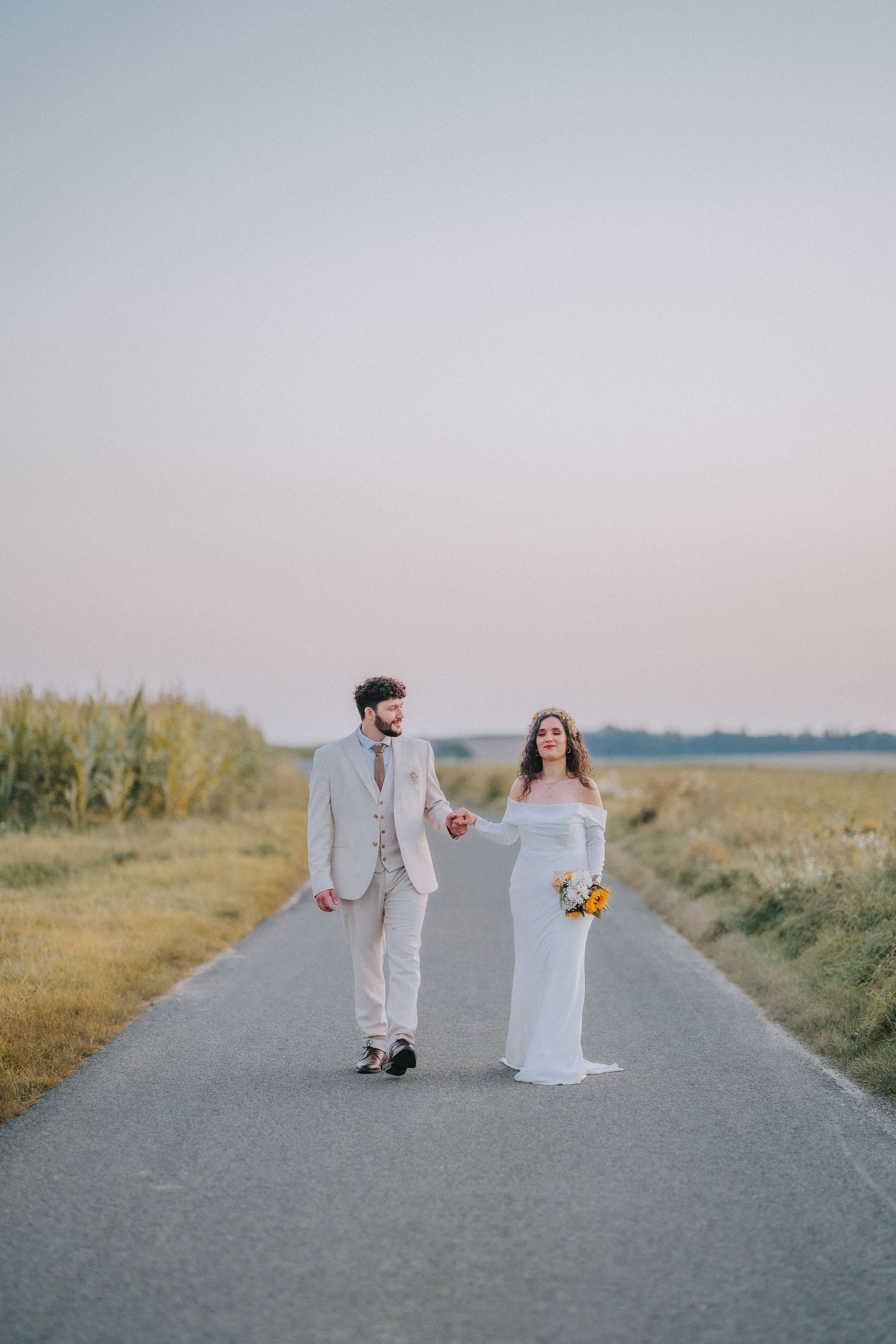 A couple walking hand in hand on a rural road at sunset, wearing elegant wedding attire and holding a sunflower bouquet.