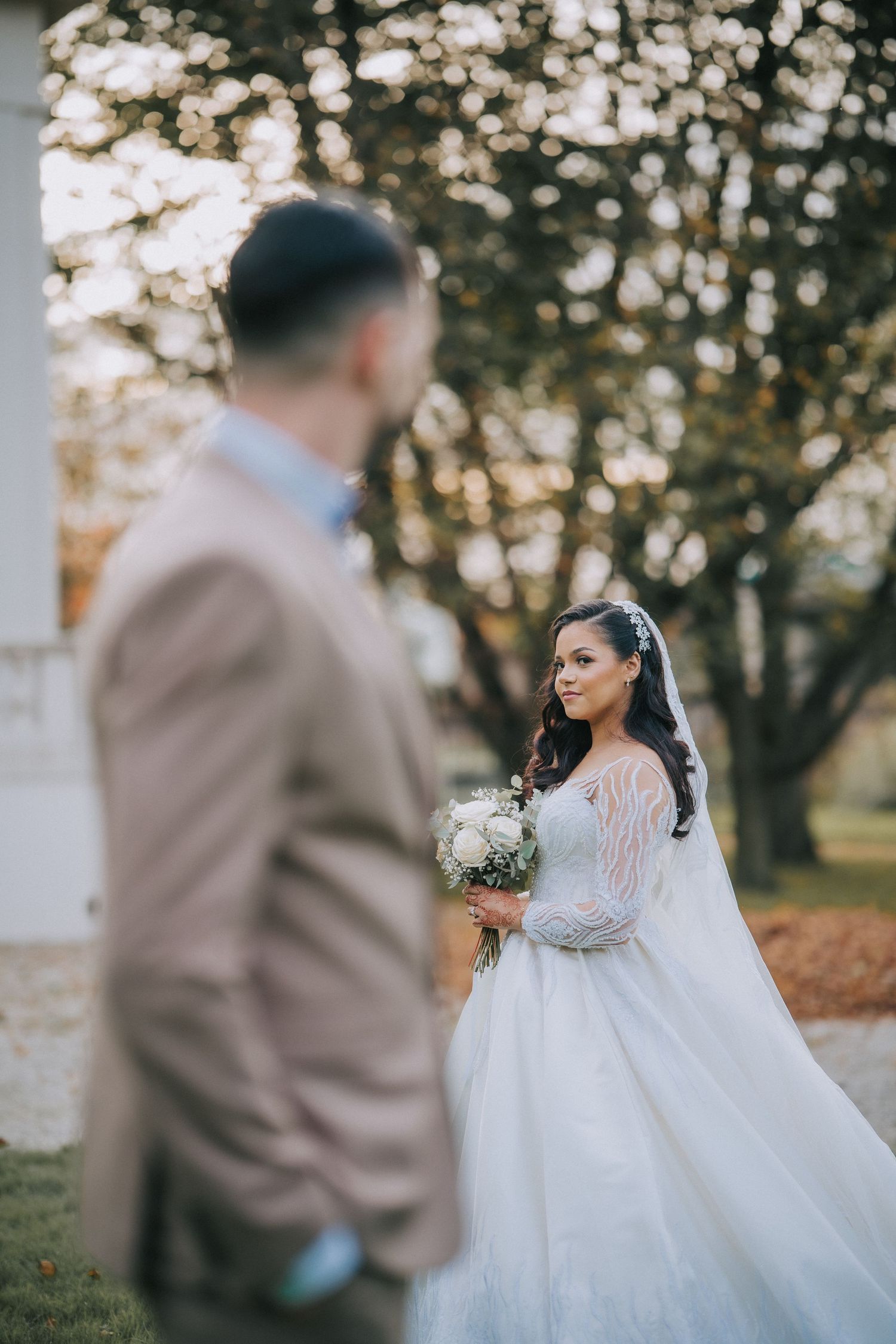 A bride in a flowing white lace gown holding a bouquet while standing outdoors during a romantic autumn wedding ceremony.