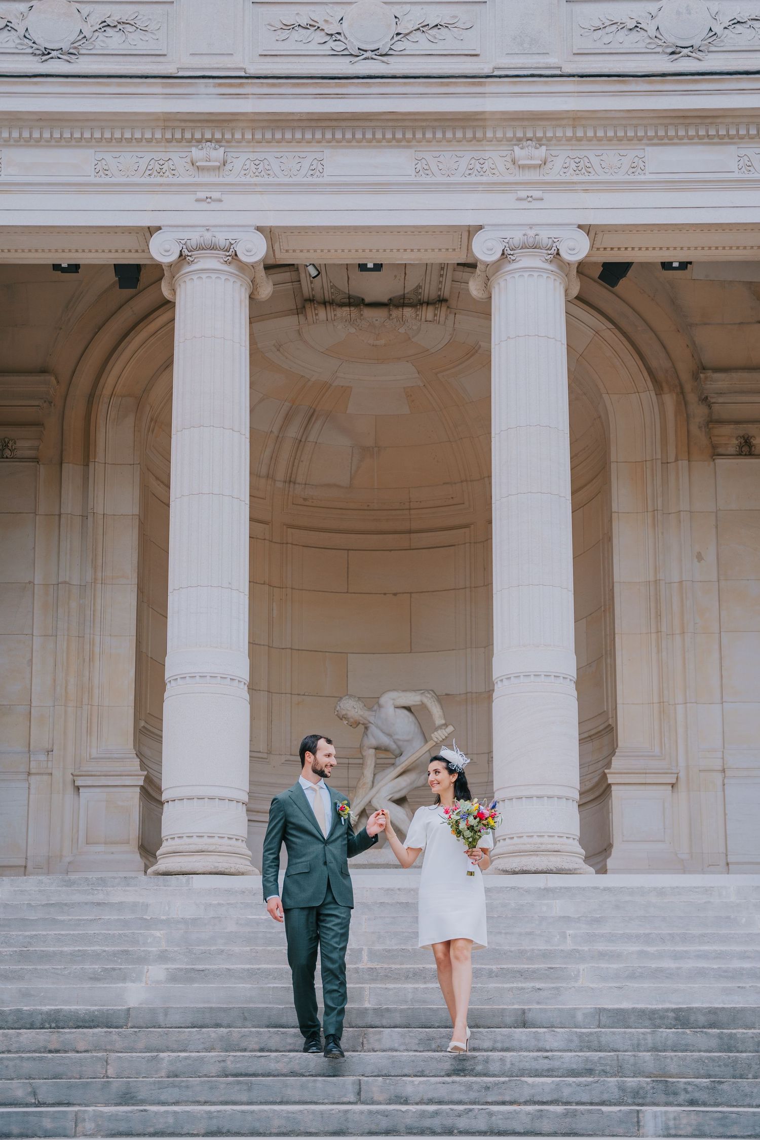 A couple in wedding attire descends elegant stone steps framed by classical white columns at a historic building.