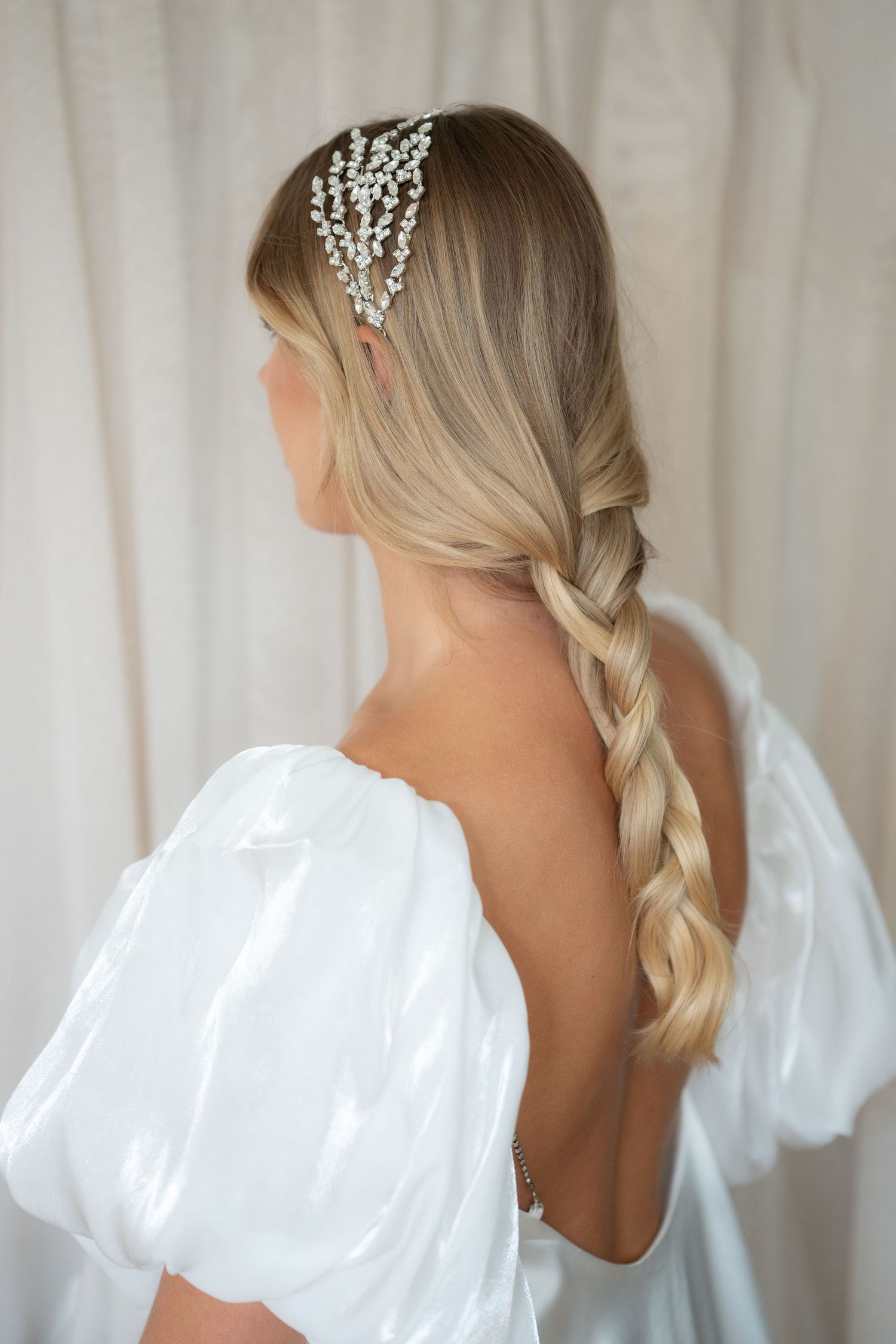 Elegant bridal hairstyle showing long blonde braid and crystal hair accessories with white backless dress.