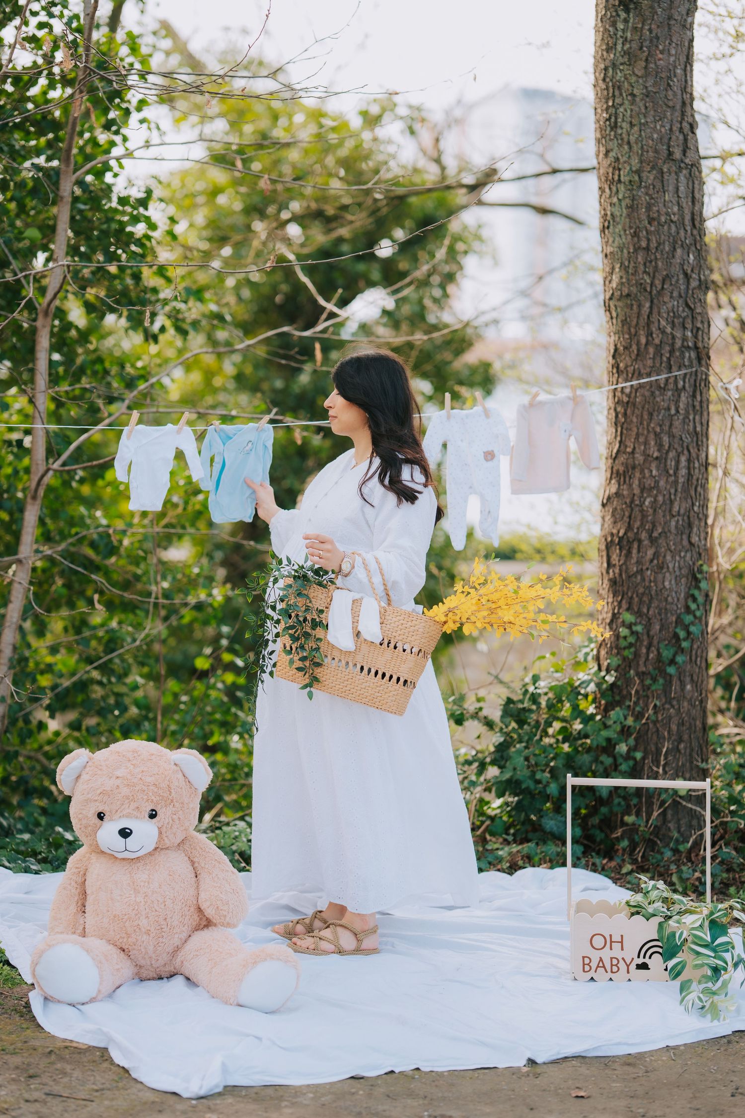 Baby clothes hanging on clothesline with teddy bear and basket in an outdoor setting.