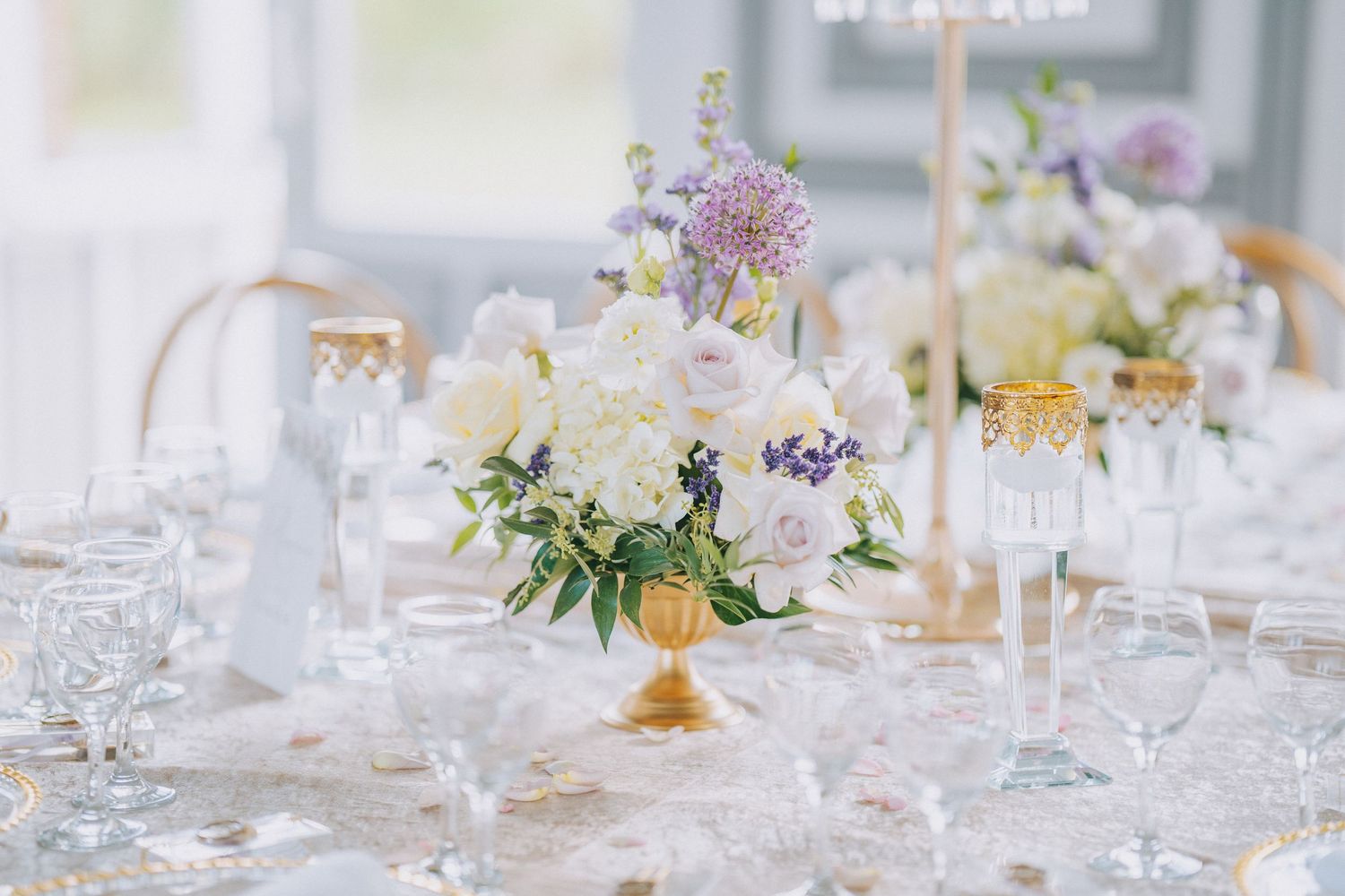 Elegant wedding table setting with pastel floral centerpiece in gold vase surrounded by crystal glassware on white tablecloth.