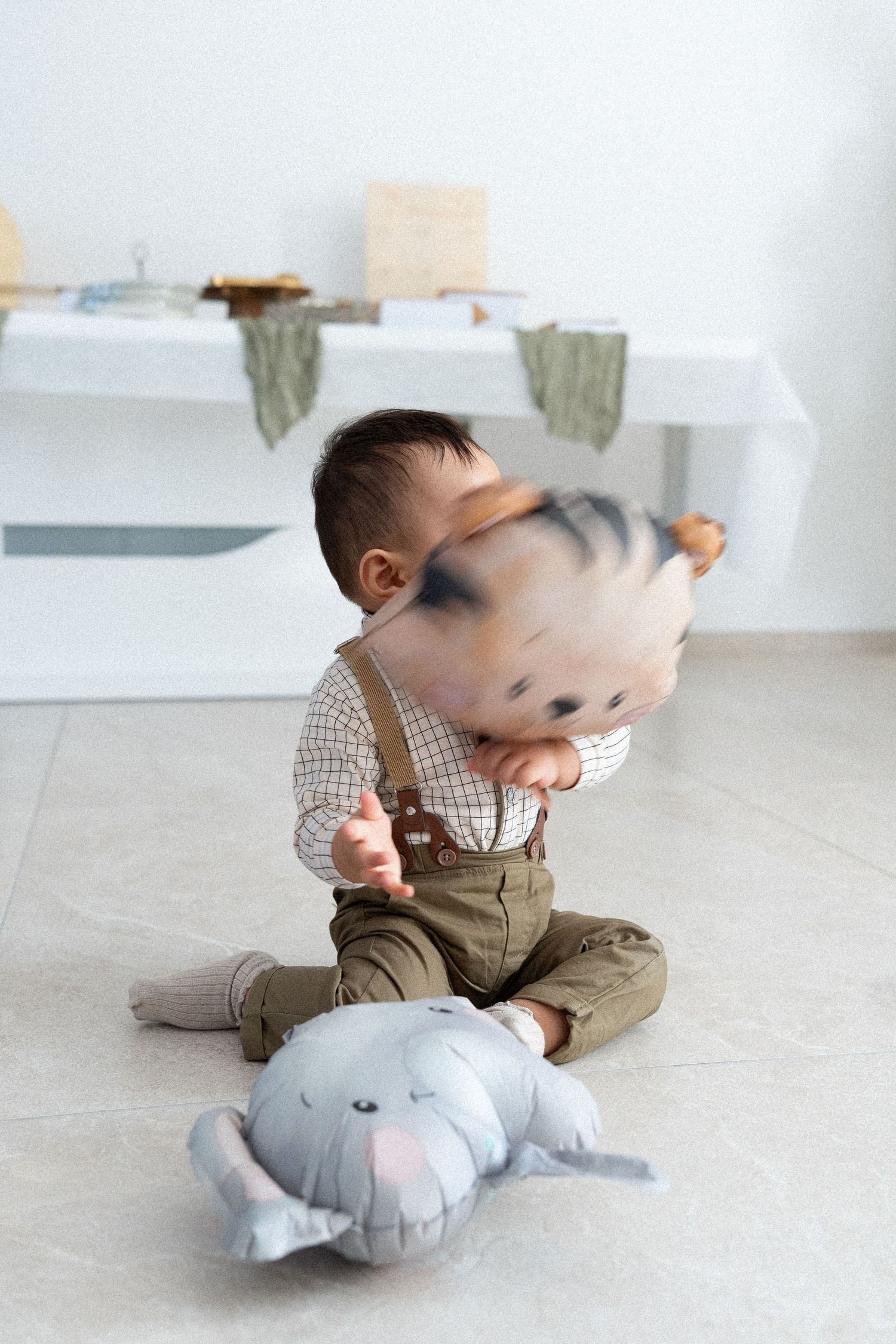 A small child in brown clothing plays with a soft gray plush elephant toy on a light-colored floor.