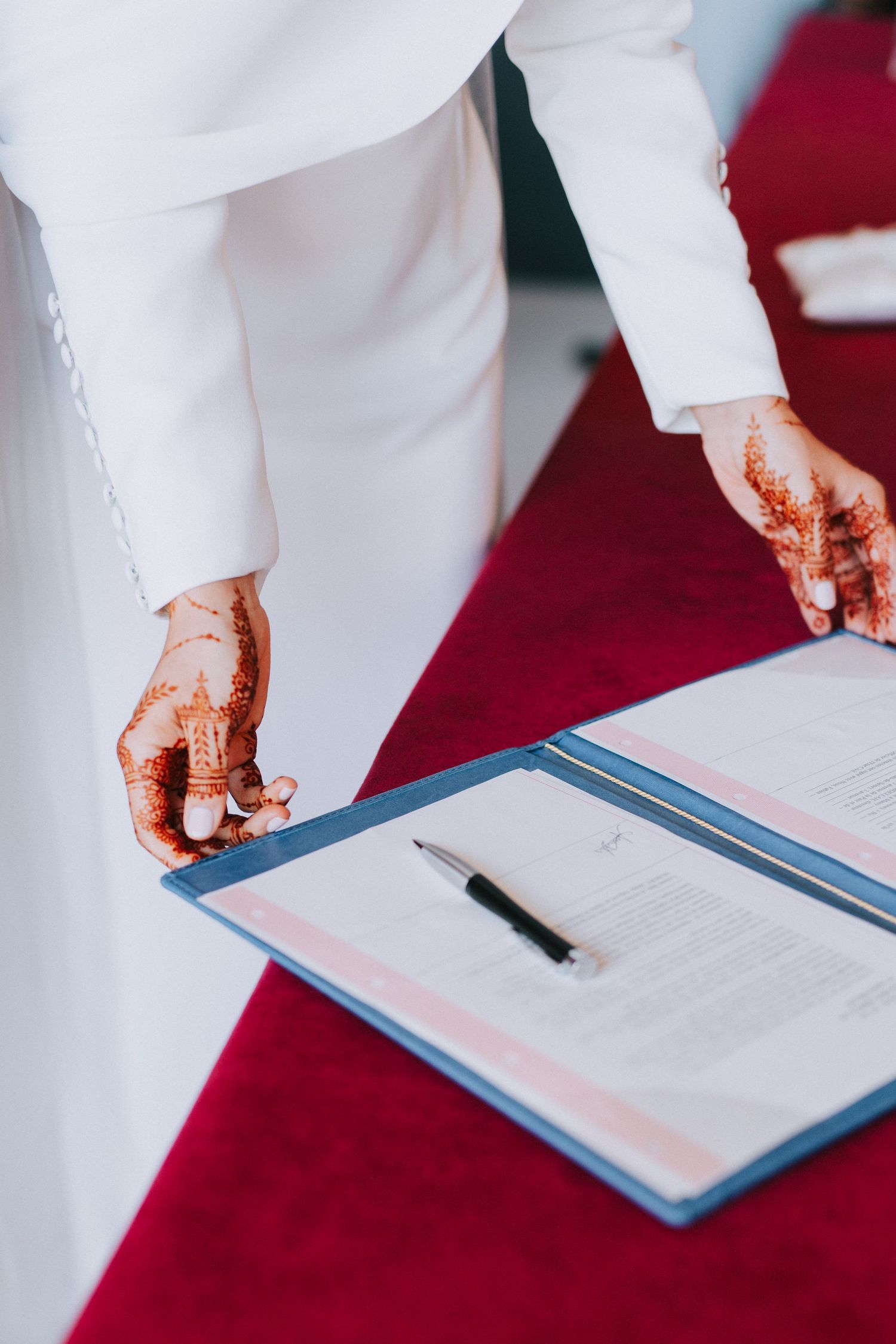 Person with henna-decorated hands signing a document on a clipboard against a red background.