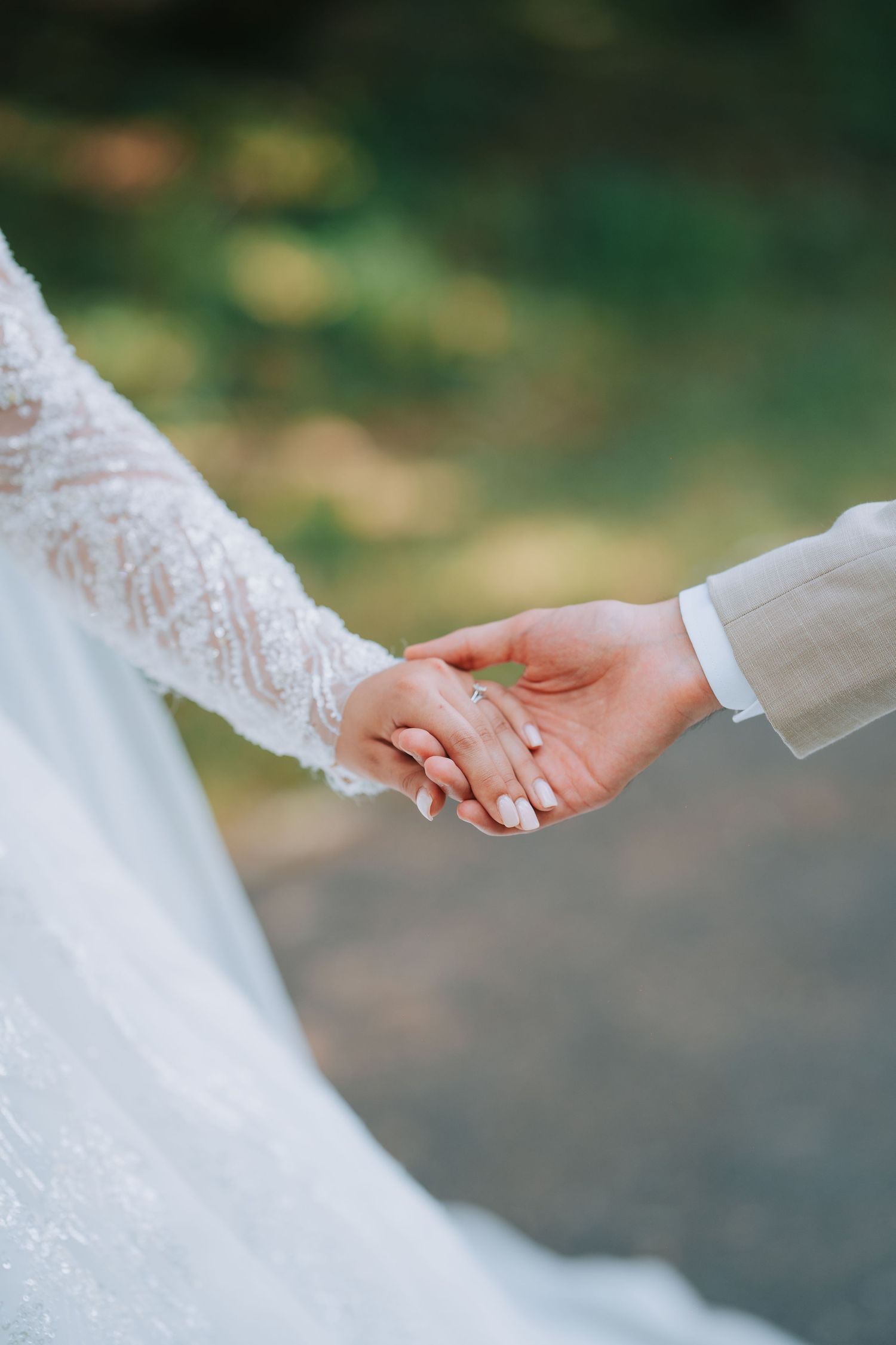 A close-up of bride and groom holding hands, showing delicate lace sleeve detail and a sparkling engagement ring.