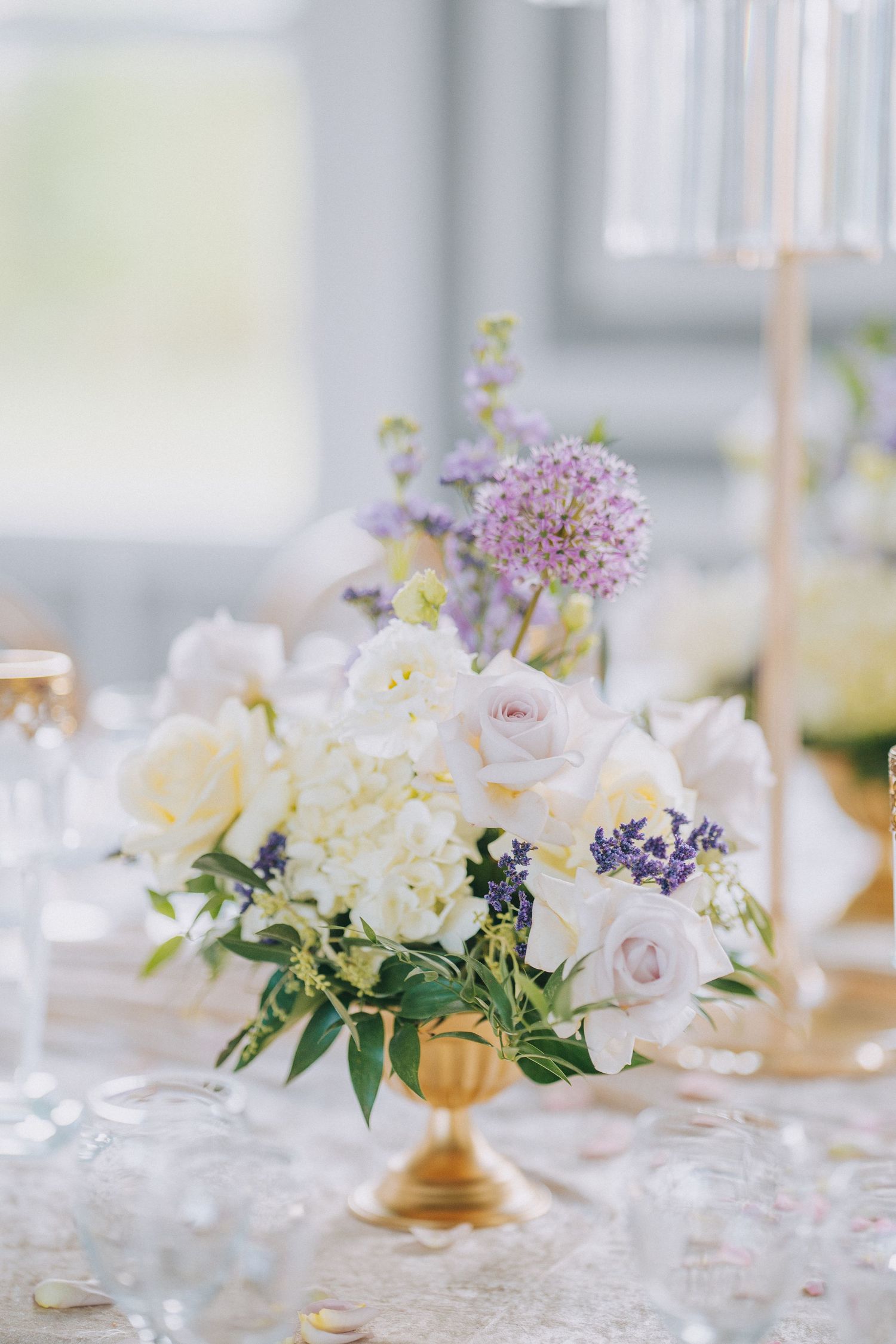 Elegant wedding centerpiece with white hydrangeas, pink roses and purple allium in a gold pedestal vase on lace tablecloth.