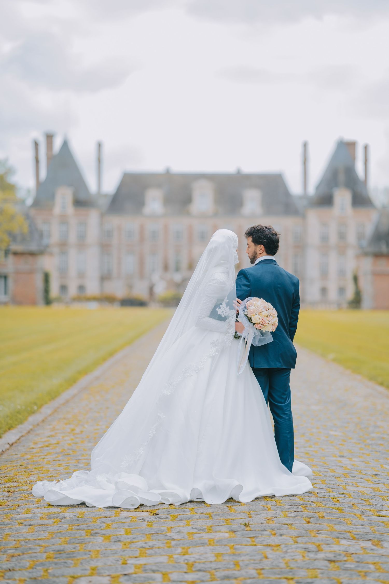 A romantic wedding couple stands on autumn leaves at an elegant French chateau with a gorgeous garden backdrop.