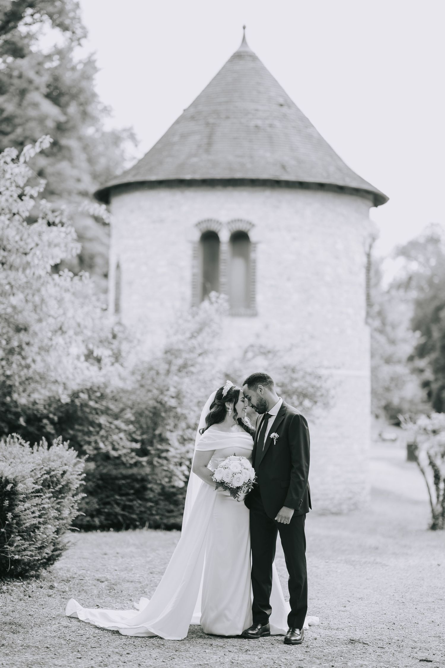 A romantic black and white photo of a couple embracing in front of a white stone church tower with a conical roof.