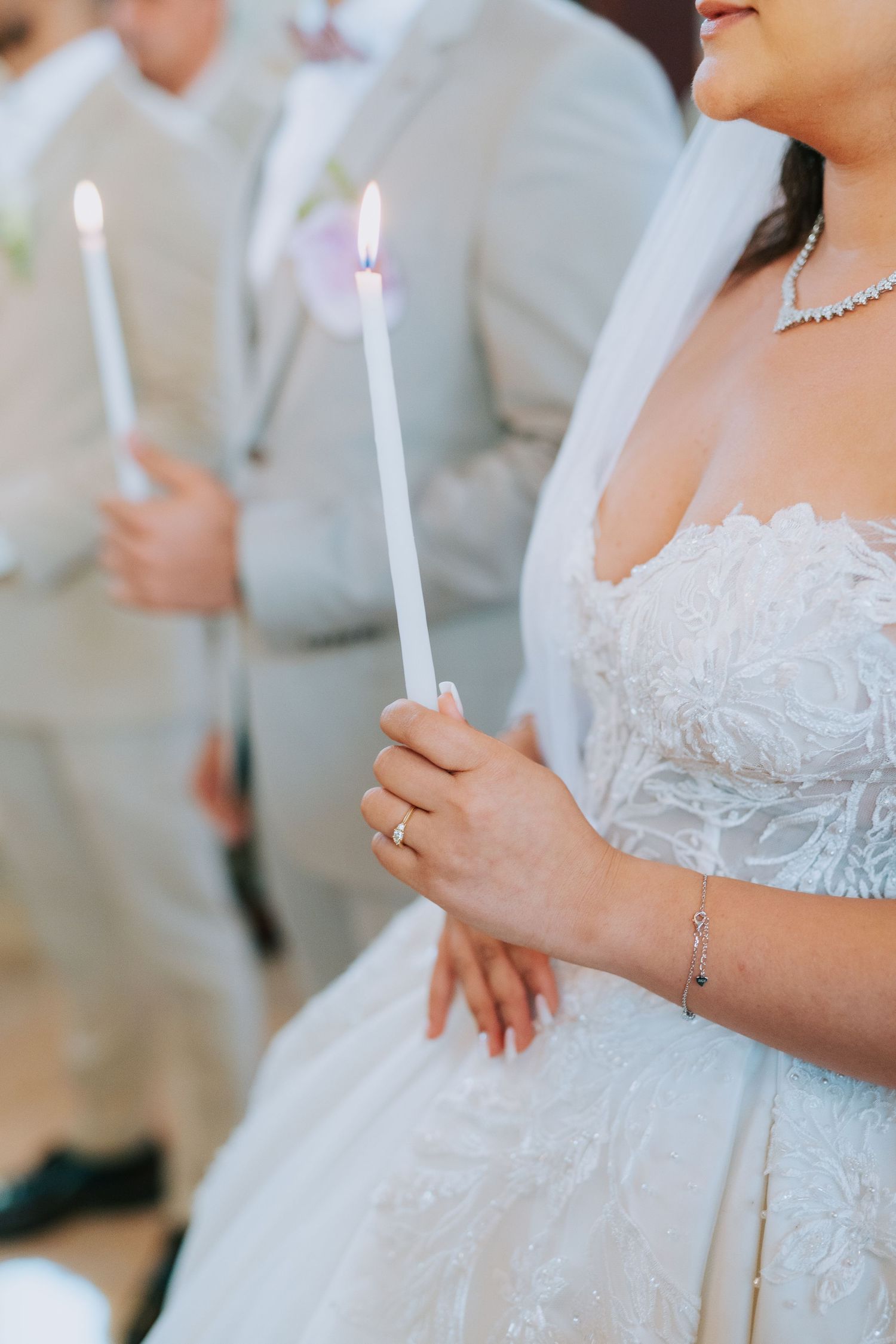 Close-up of couple holding lit white candles at wedding ceremony, with bride wearing lace dress and sparkling jewelry.