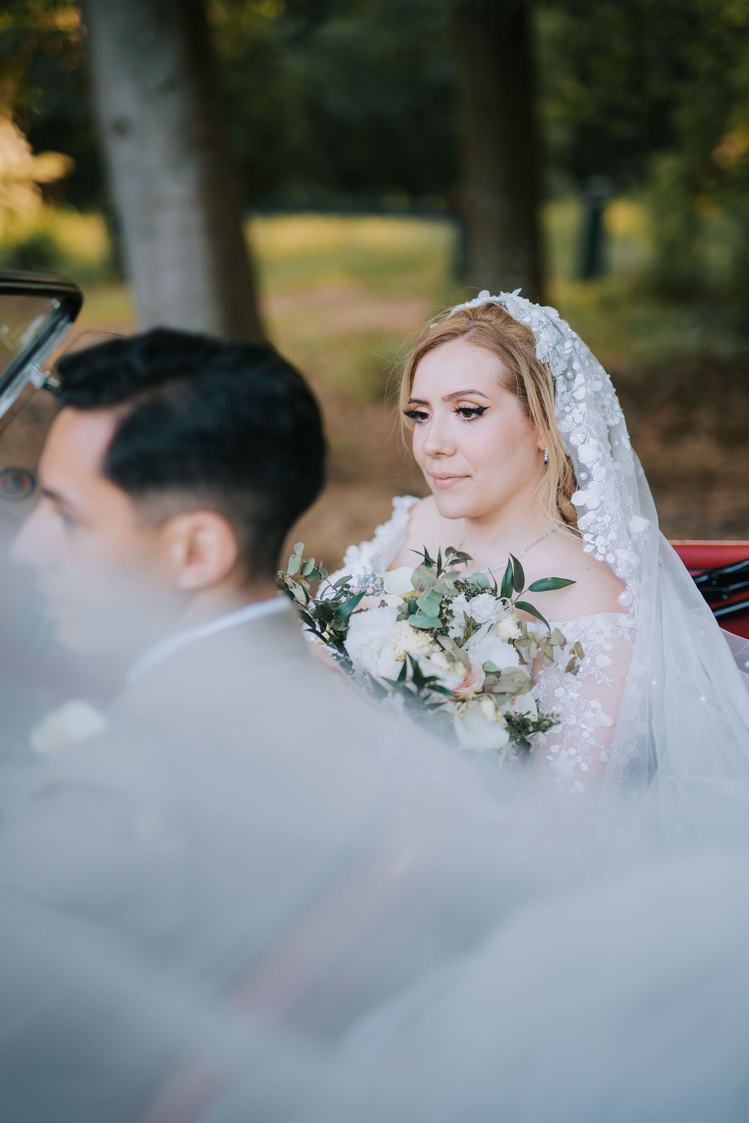 A bride in a white lace veil holds a soft pastel wedding bouquet while sitting in a car on her wedding day.
