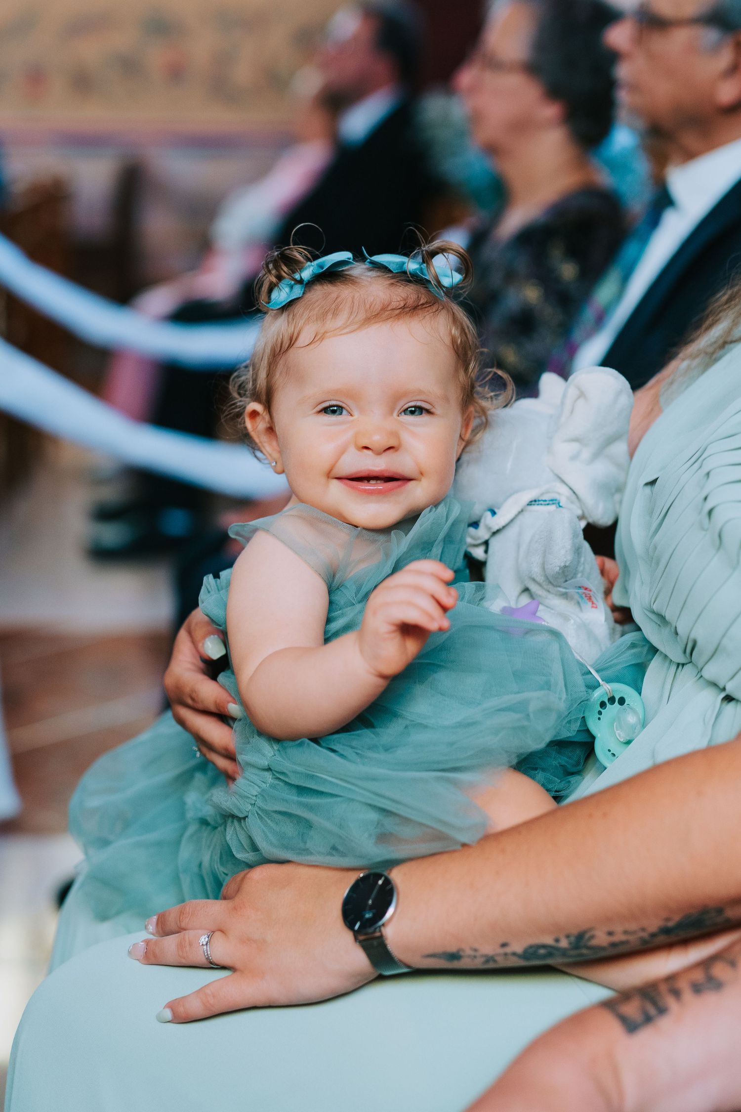 A smiling baby in a turquoise dress with small blue bows in their hair sits on someone's lap at an event.