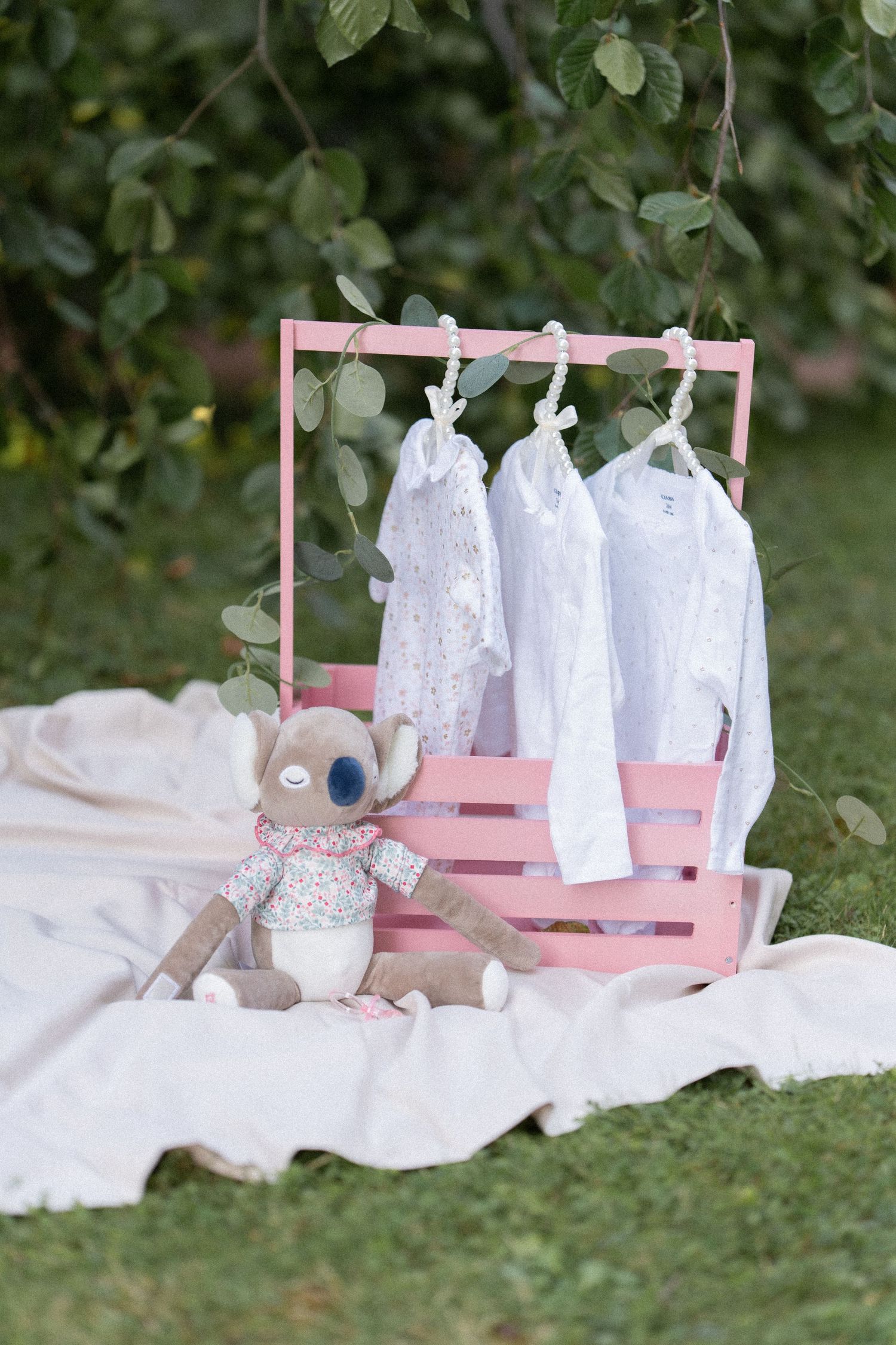 Pink wooden display rack with white baby dresses hanging and a plush teddy bear sitting beside it on a blanket outdoors.