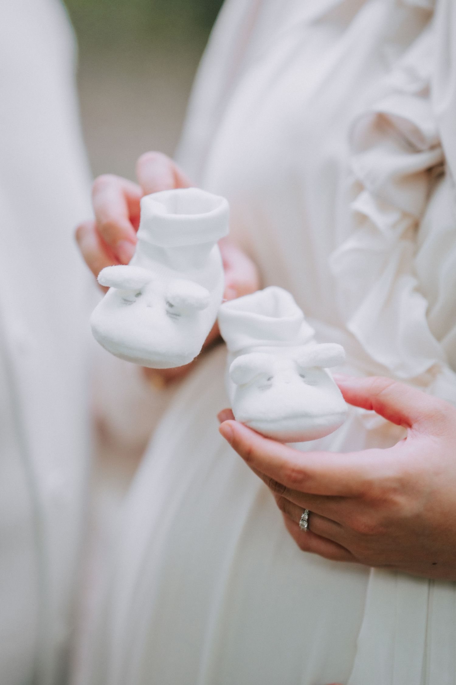 White baby booties held gently against flowing fabric with a wedding ring visible.