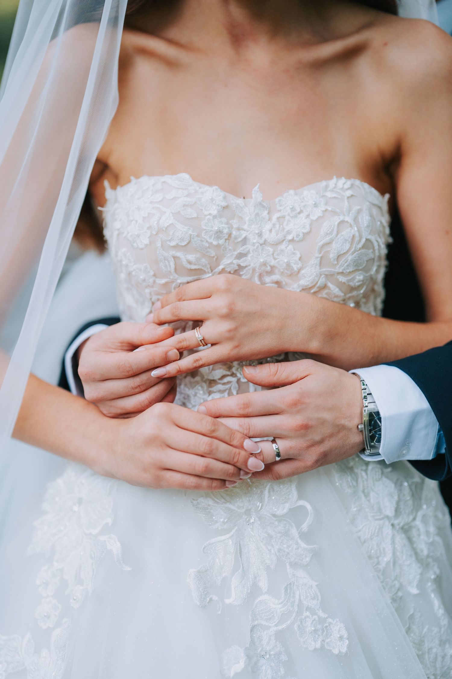 Close-up of newlywed couple's hands embracing, with bride wearing detailed lace wedding dress and sparkling ring.
