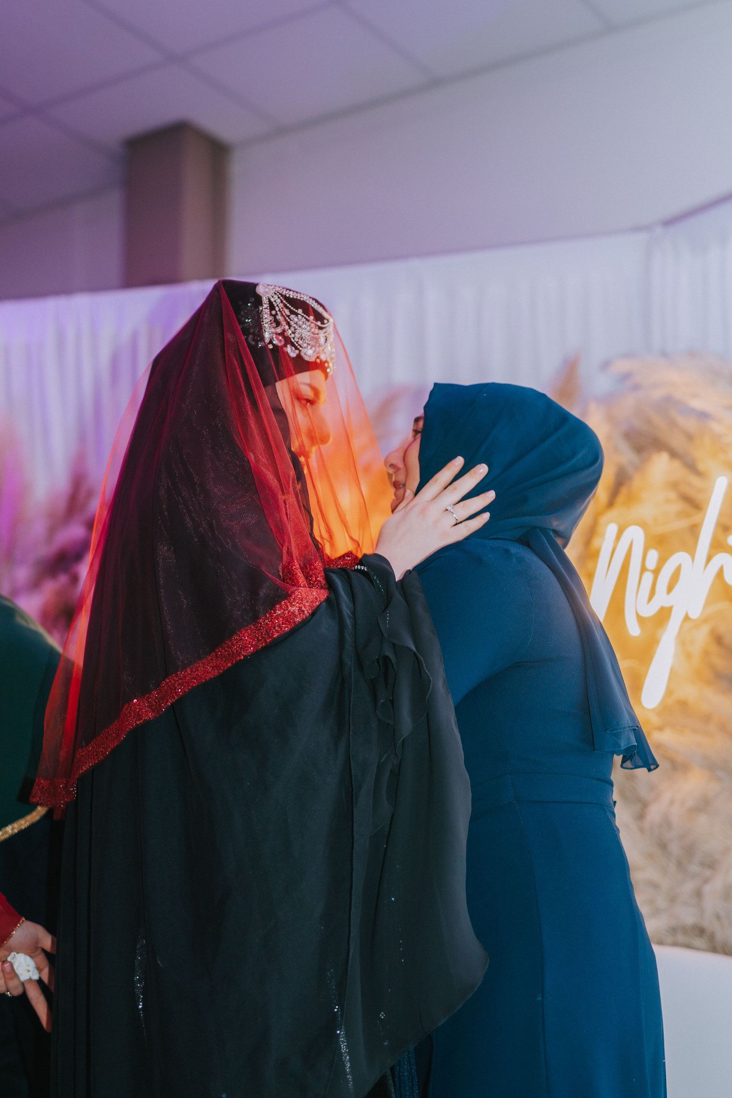 Two women in traditional hijabs embracing at celebratory event with neon sign in background.