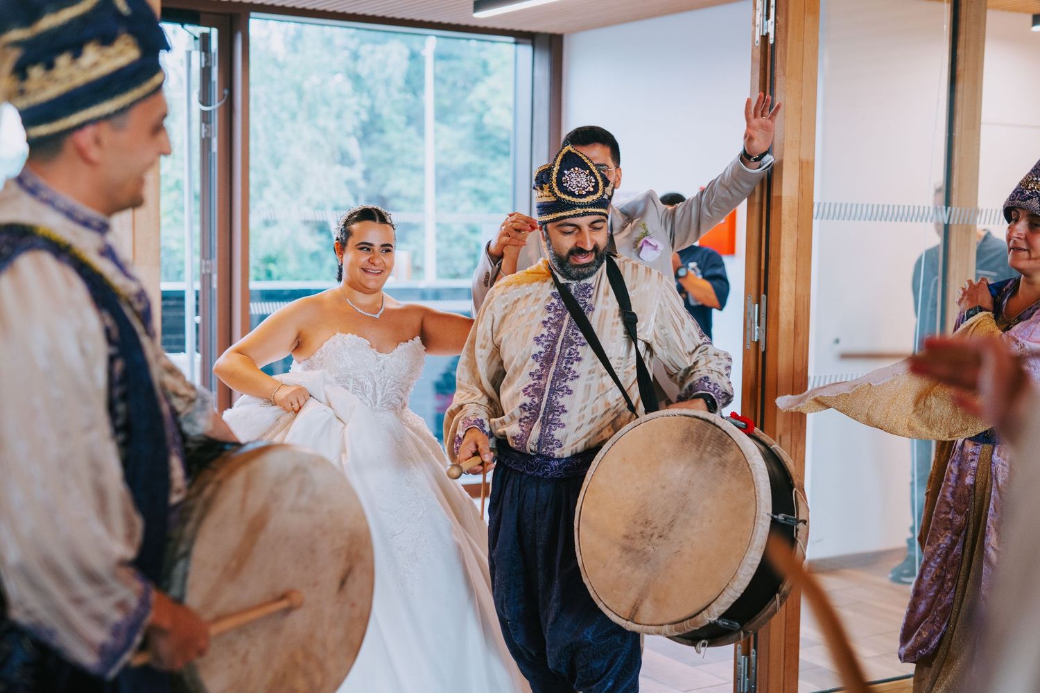 Traditional musicians with drums entertain a person in a white dress at an indoor celebration event.