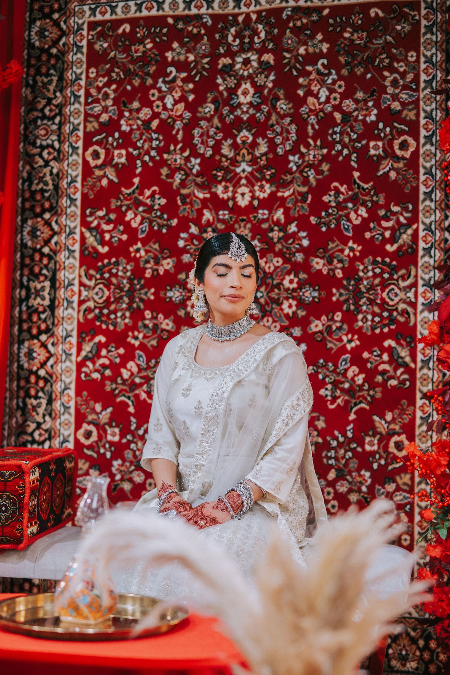 Traditional bridal portrait against red patterned tapestry with white attire and jewelry.