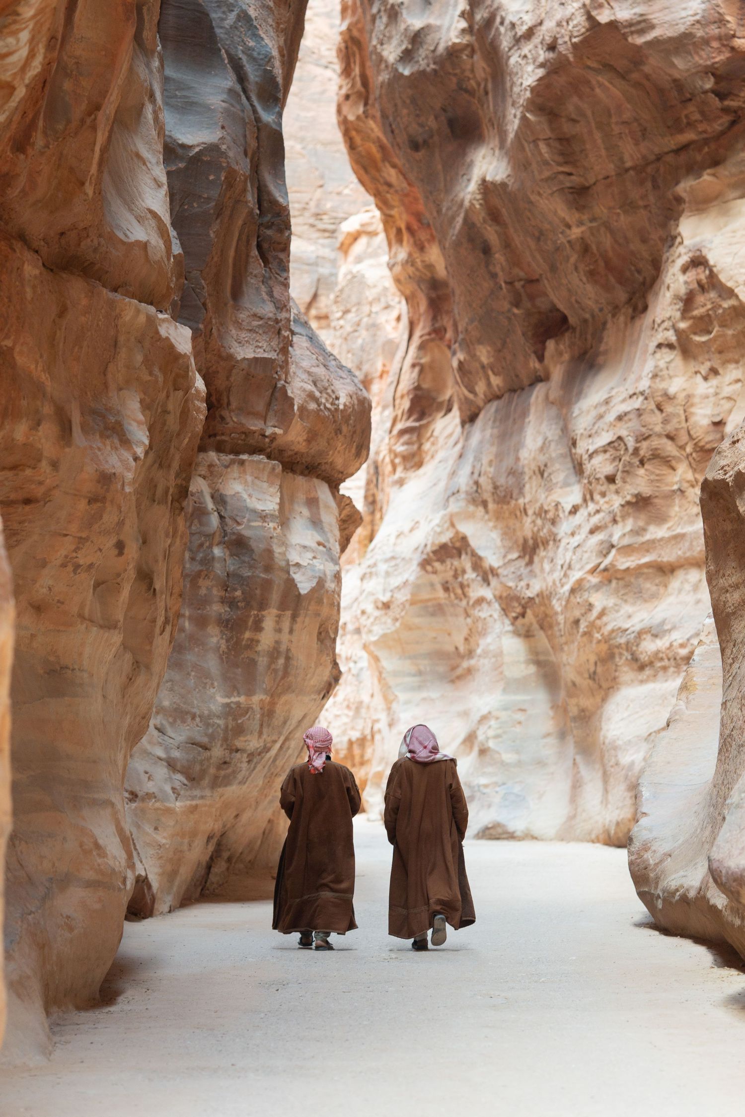 Two figures in traditional robes walking through narrow desert canyon pathway.