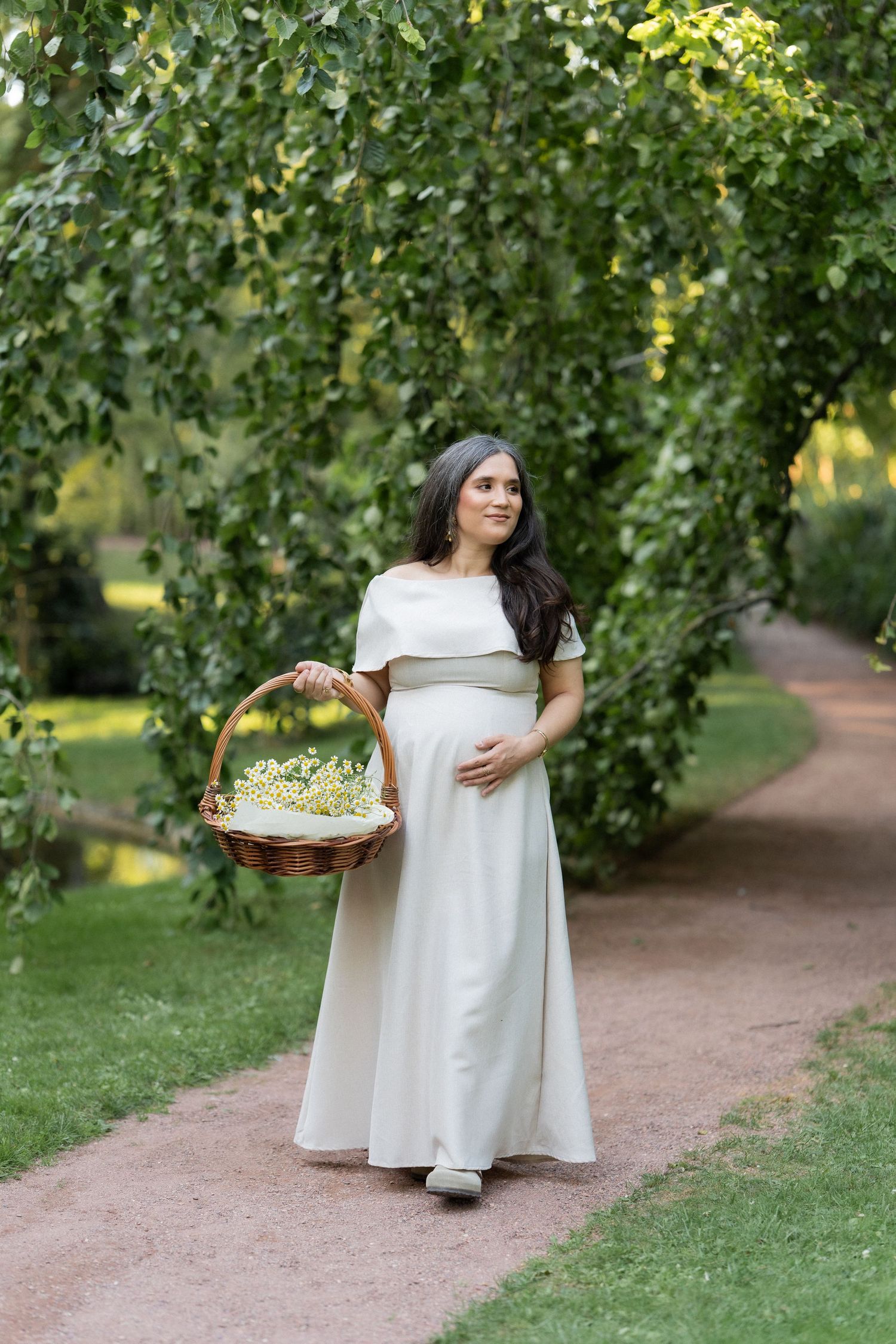 A person in a flowing white dress walks along a garden path carrying a wicker basket under green leafy trees.
