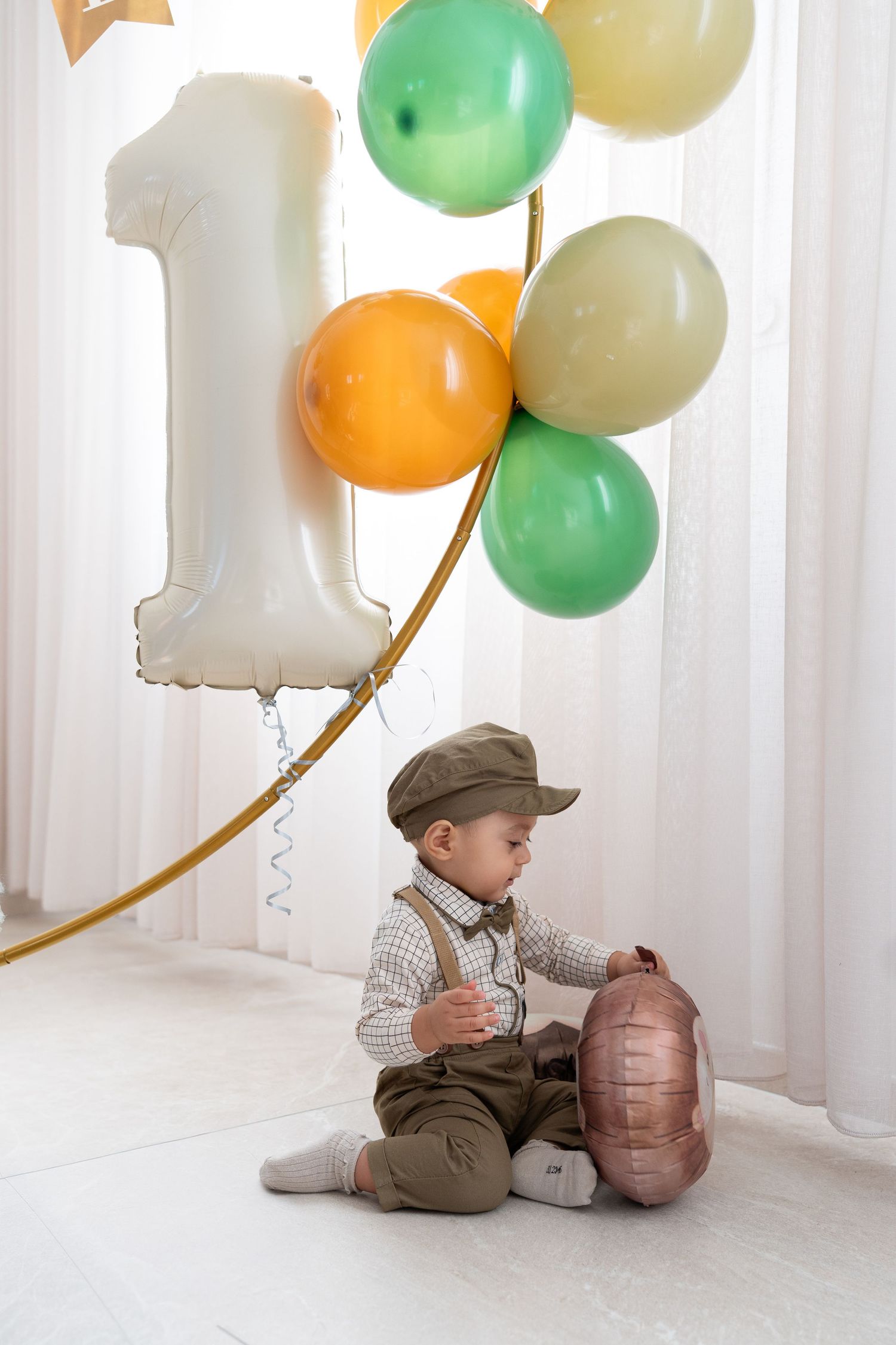 Baby in vintage-style outfit sits with decorative balloons and a brown suitcase against white backdrop.