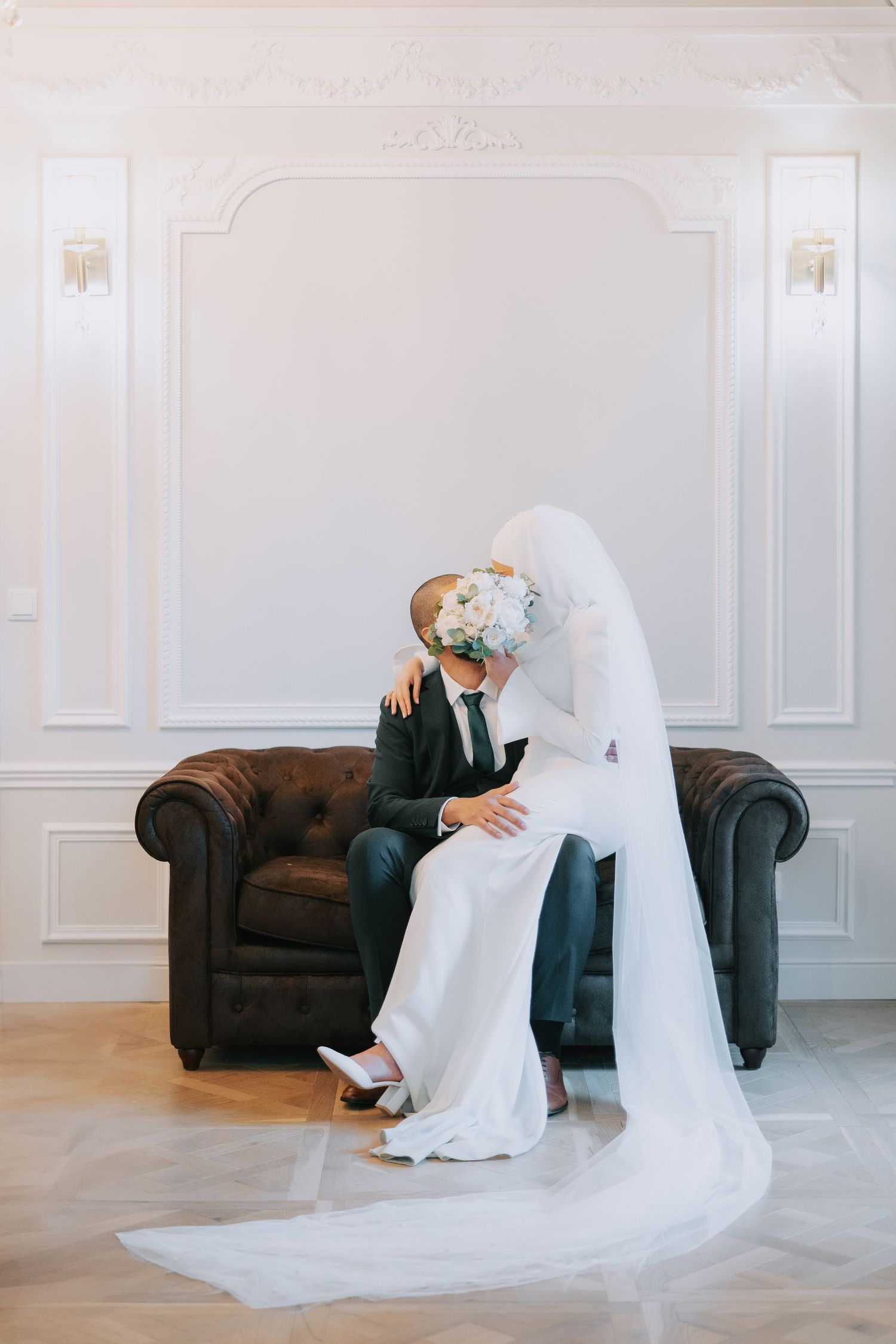 A wedding couple sits together on a luxurious black leather chesterfield sofa in an elegant white room.
