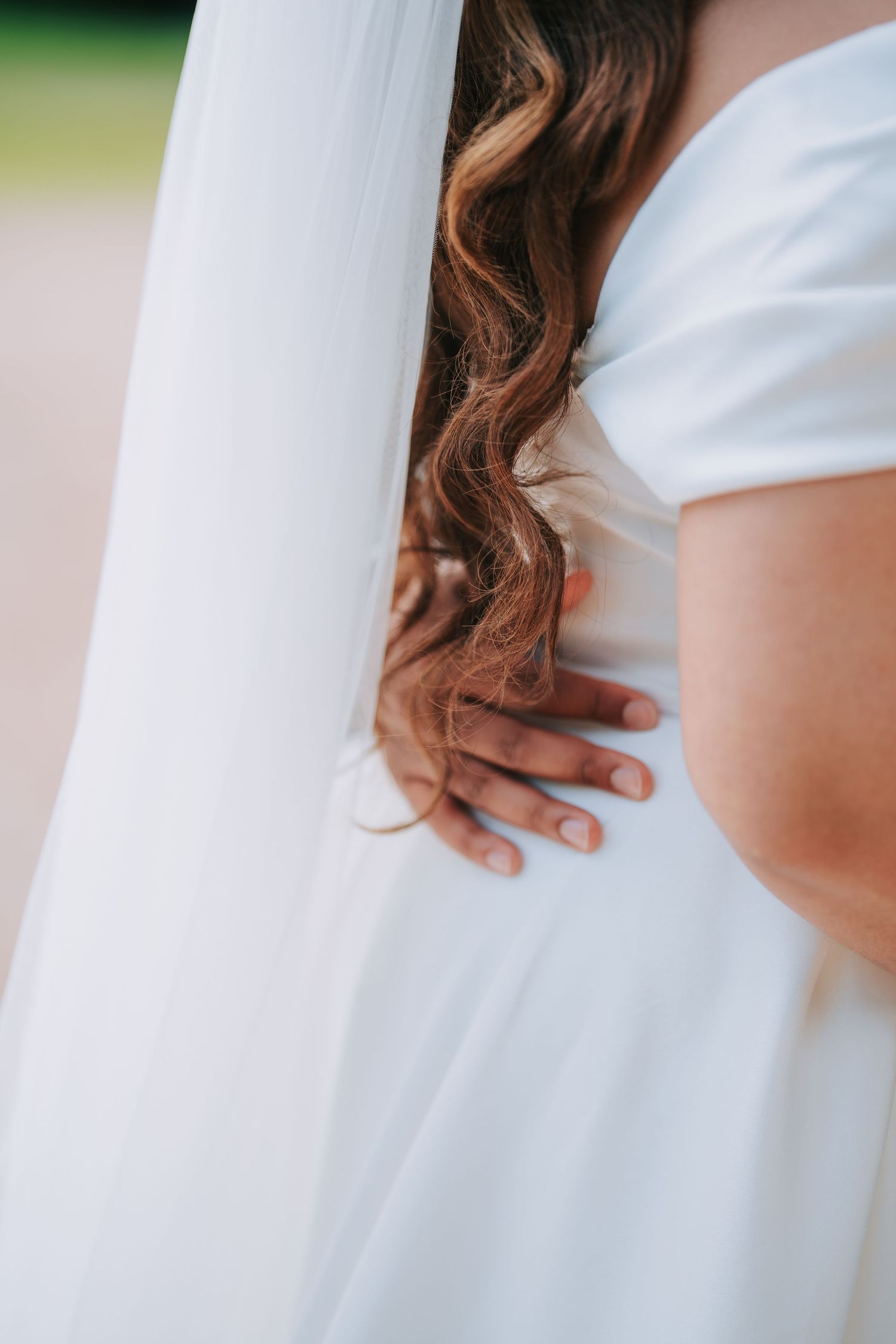 Close-up detail of wavy brown hair and hand resting against a white wedding dress fabric.