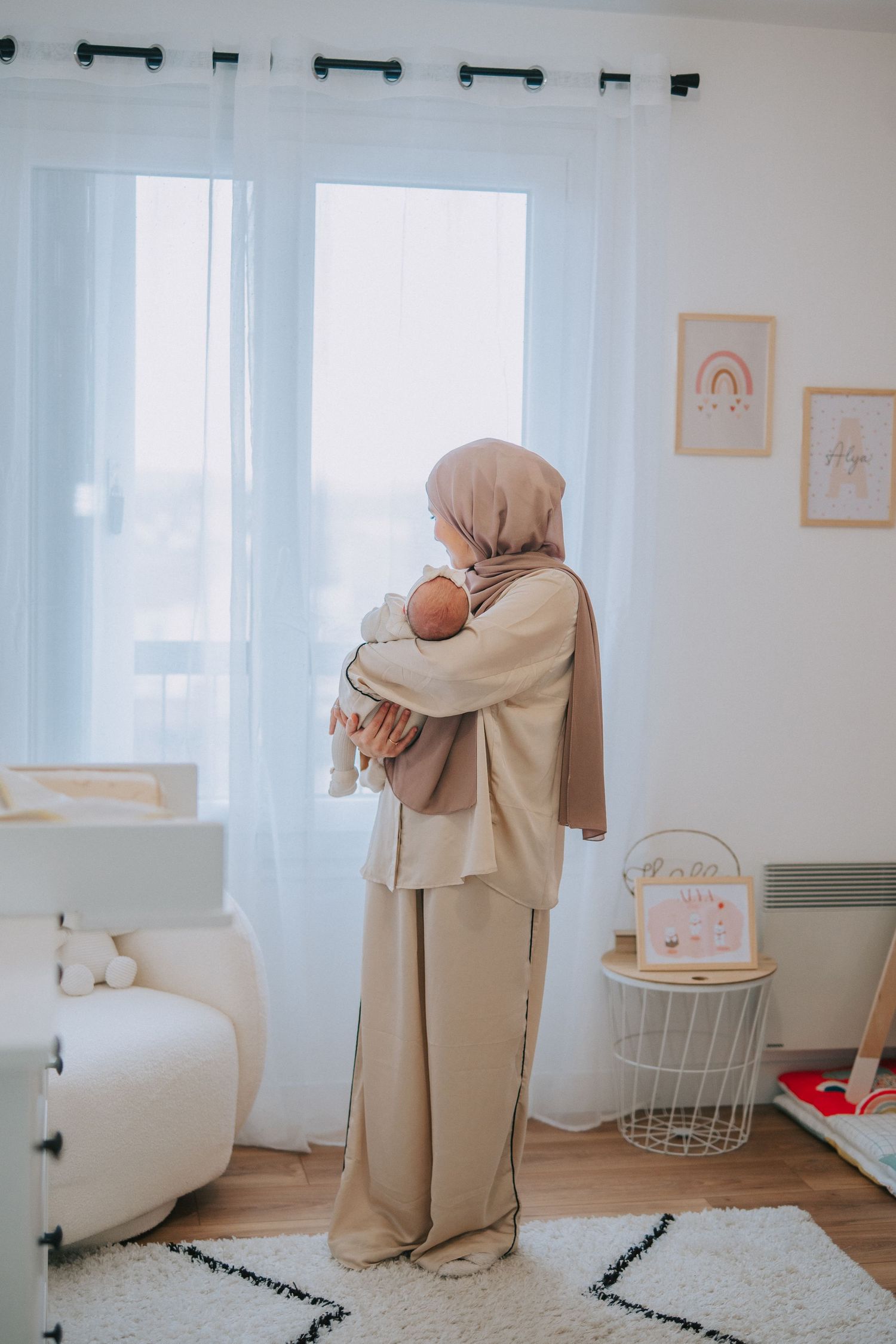 Person in beige outfit standing by window holding baby in bright nursery room.