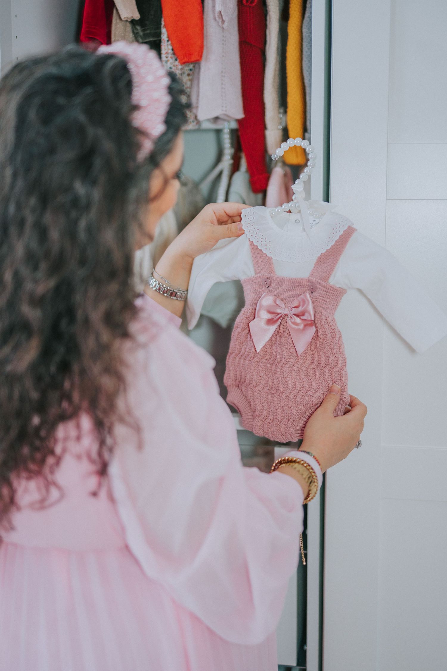 Pink baby romper with bow being held up against a closet of colorful clothing.