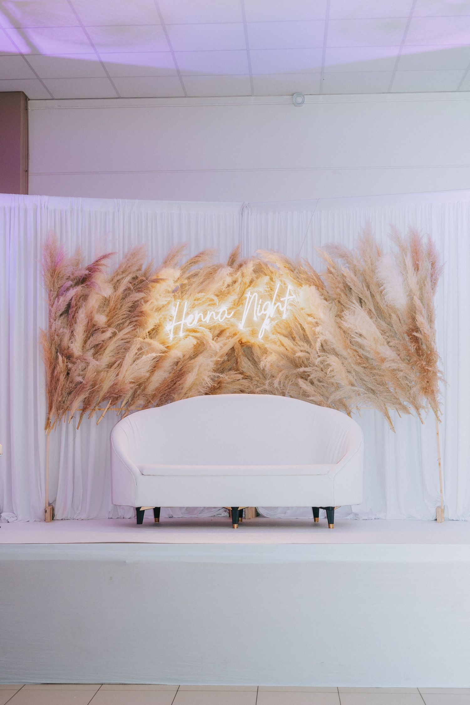 Elegant wedding stage setup with white sofa and dried pampas grass decor featuring neon 'forever love' sign.