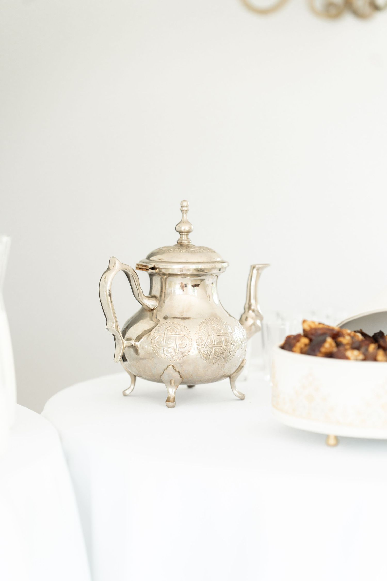 Vintage silver teapot with ornate details sits elegantly on white surface next to serving dish.