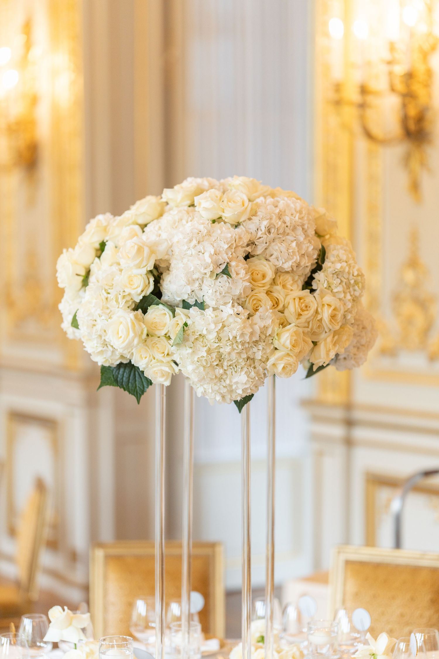 Elegant tall white rose and hydrangea floral centerpiece on glass stand in luxurious wedding reception venue.