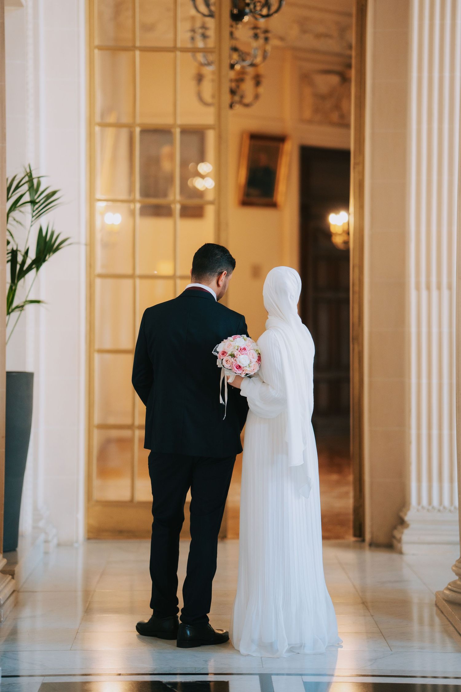 A newly married couple in formal attire stand in an elegant hallway with white columns and a pink floral bouquet.