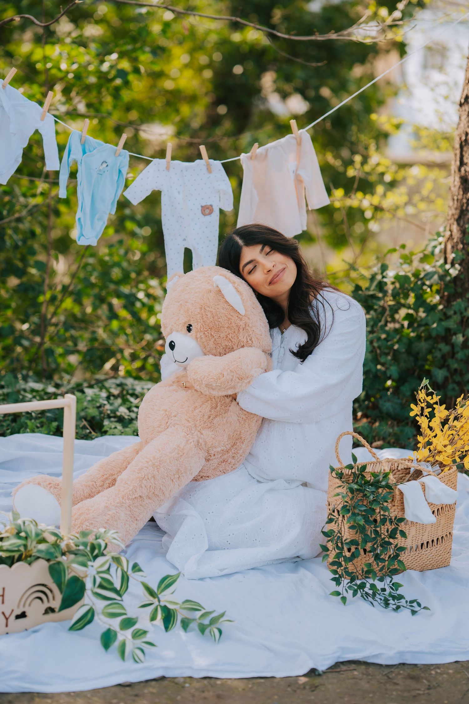 Maternity photo setup with baby clothes on clothesline and large teddy bear on white blanket in garden setting.