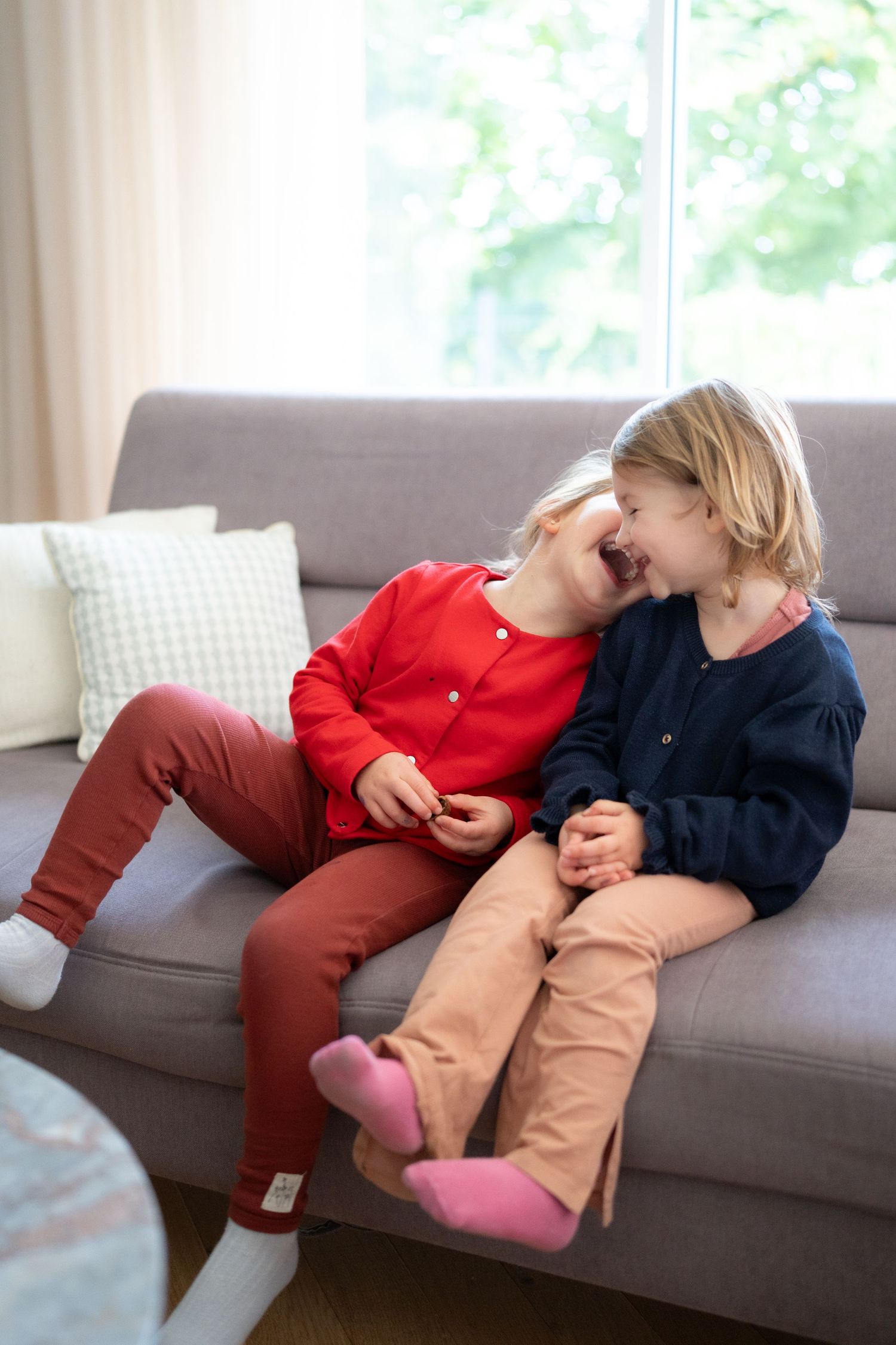 Two children laughing together on gray couch by window.