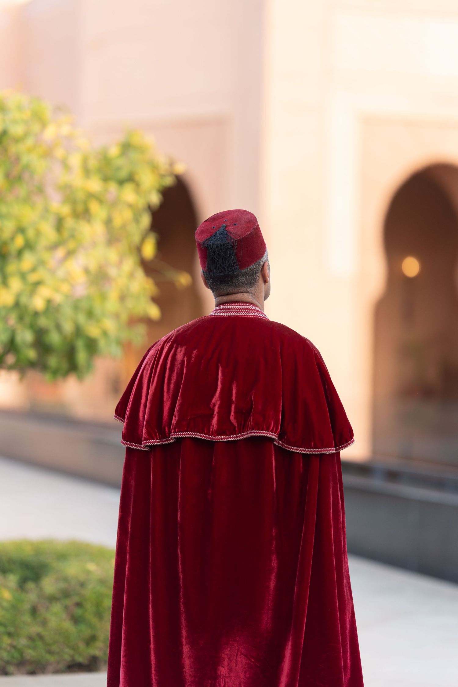 Figure wearing traditional red velvet cape and fez hat walking near arched colonnade.