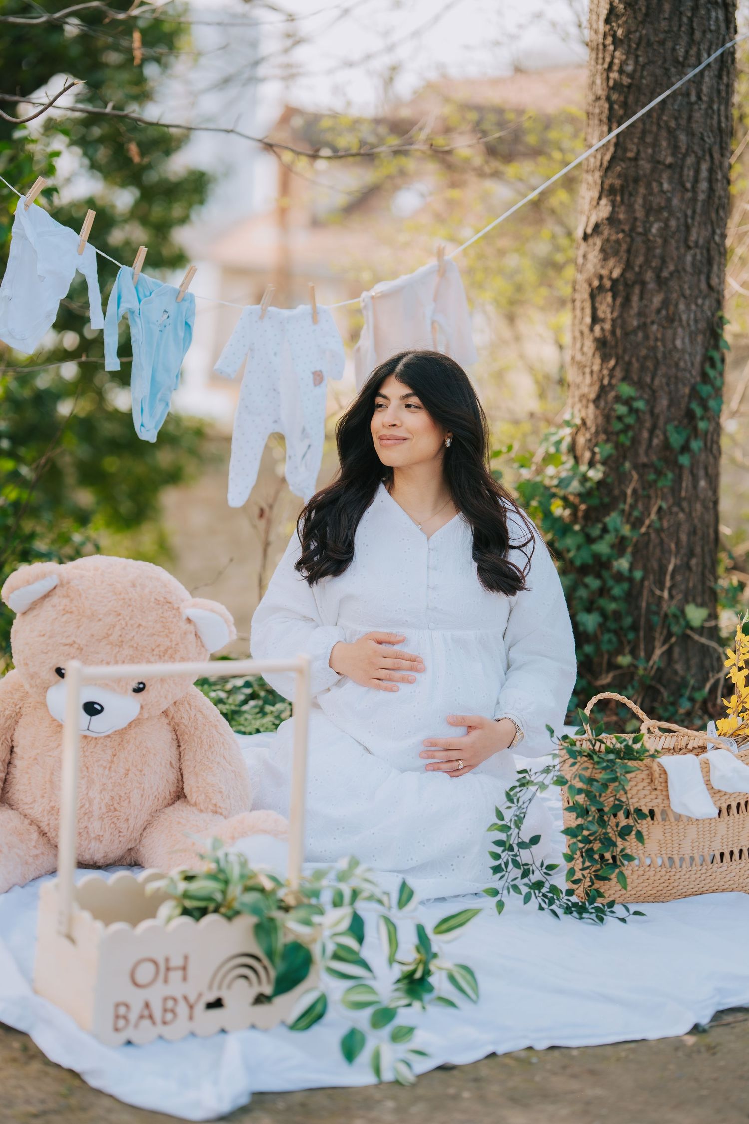 Pregnancy photoshoot with baby clothes hanging on line and teddy bear in garden setting.