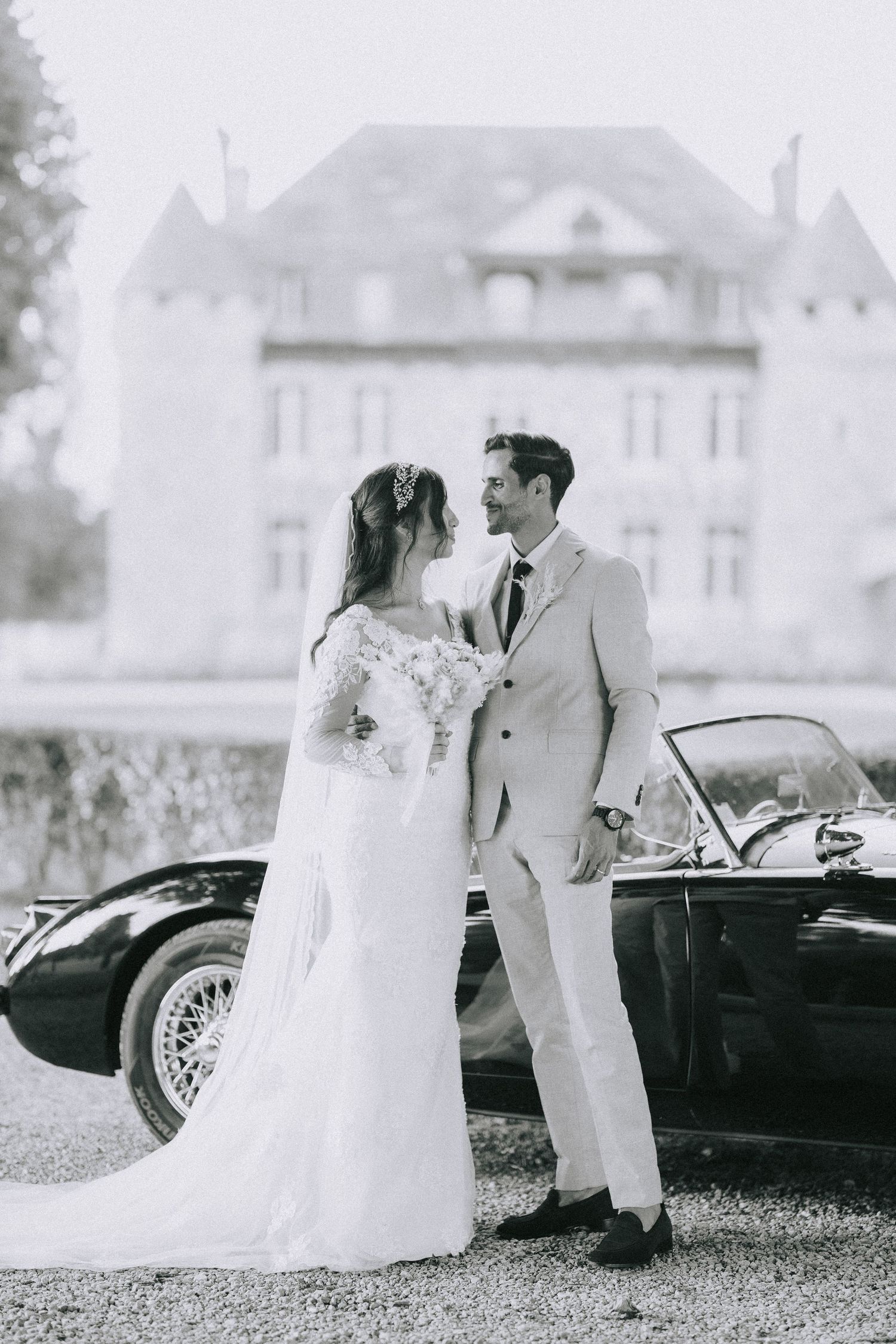 Black and white wedding photo of a couple posing next to a vintage car with a grand mansion in the background.