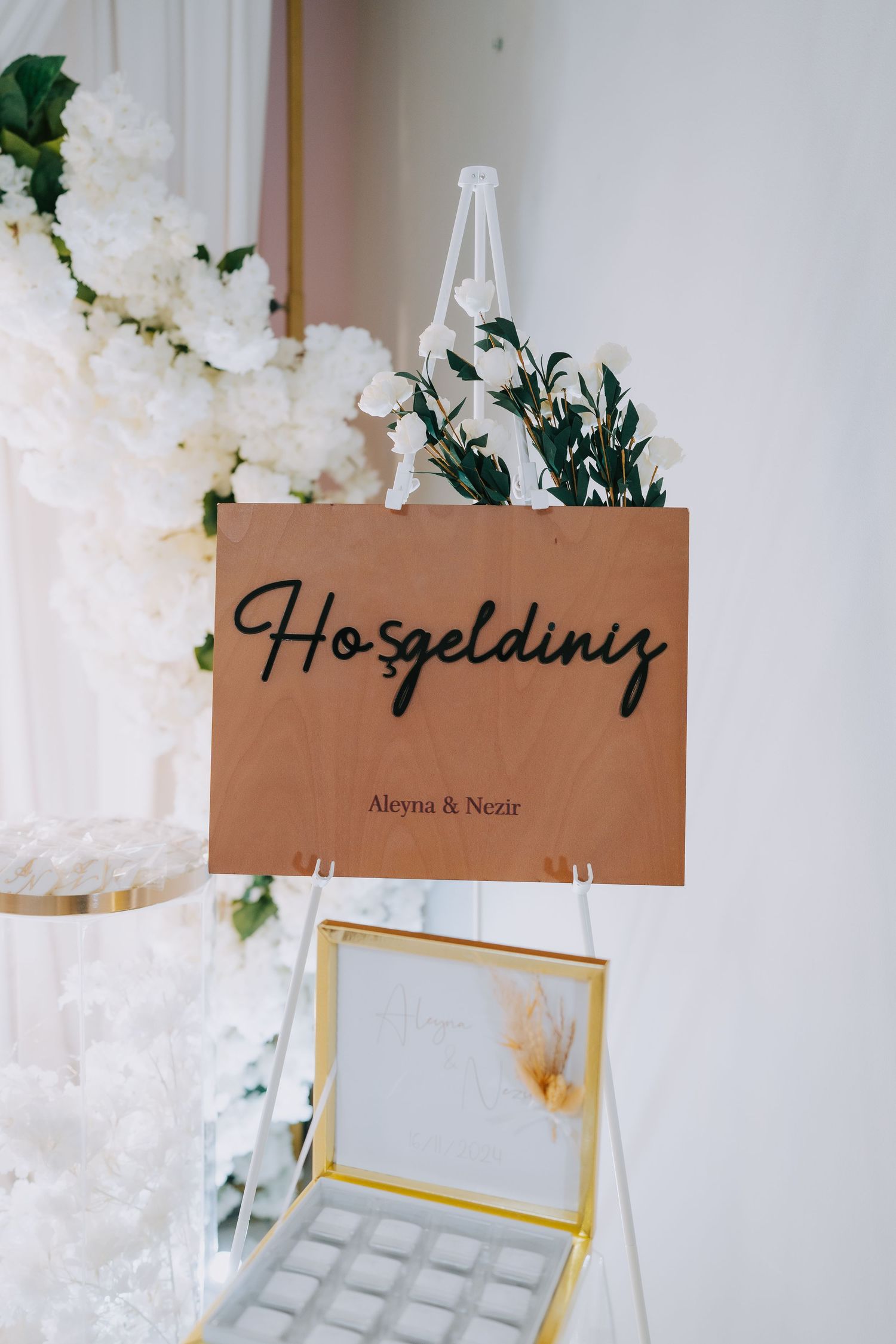 Brown welcome sign with 'Hosgeldiniz' text decorated with greenery and white flowers.