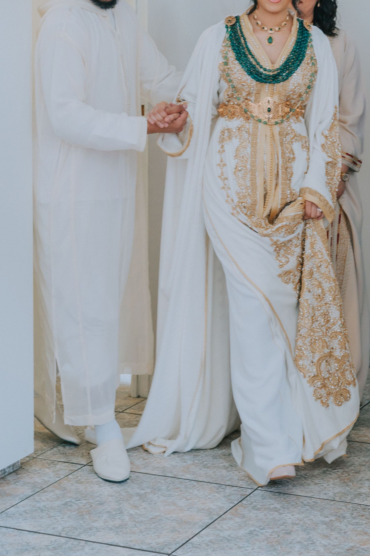 Traditional Moroccan couple in white and gold wedding attire holding hands.