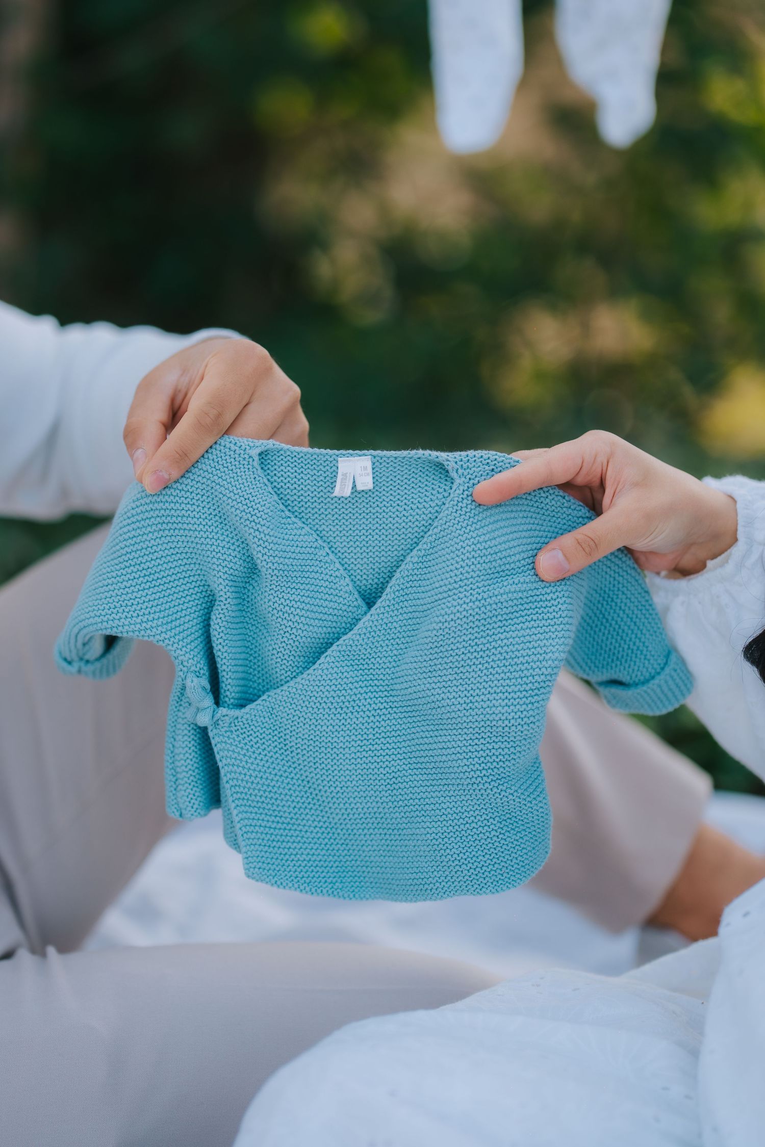 A small turquoise blue baby sweater being held up against a blurred outdoor background.