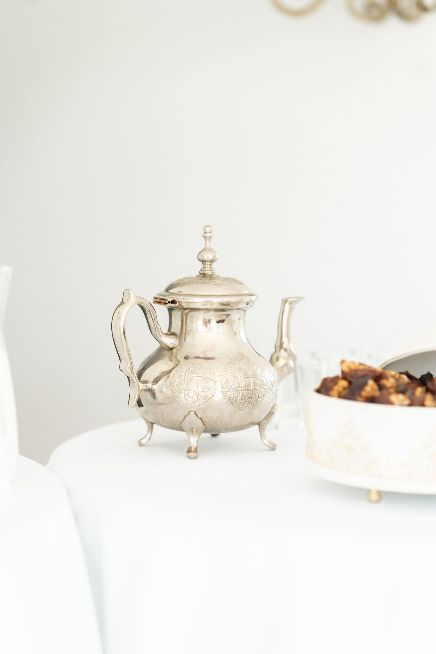Vintage silver teapot with ornate details sits elegantly on white table next to serving dish.