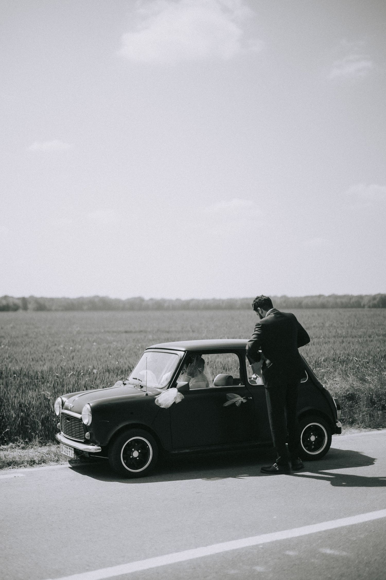 A black and white photo shows a classic Mini Cooper parked on a rural road with someone sitting on its roof.