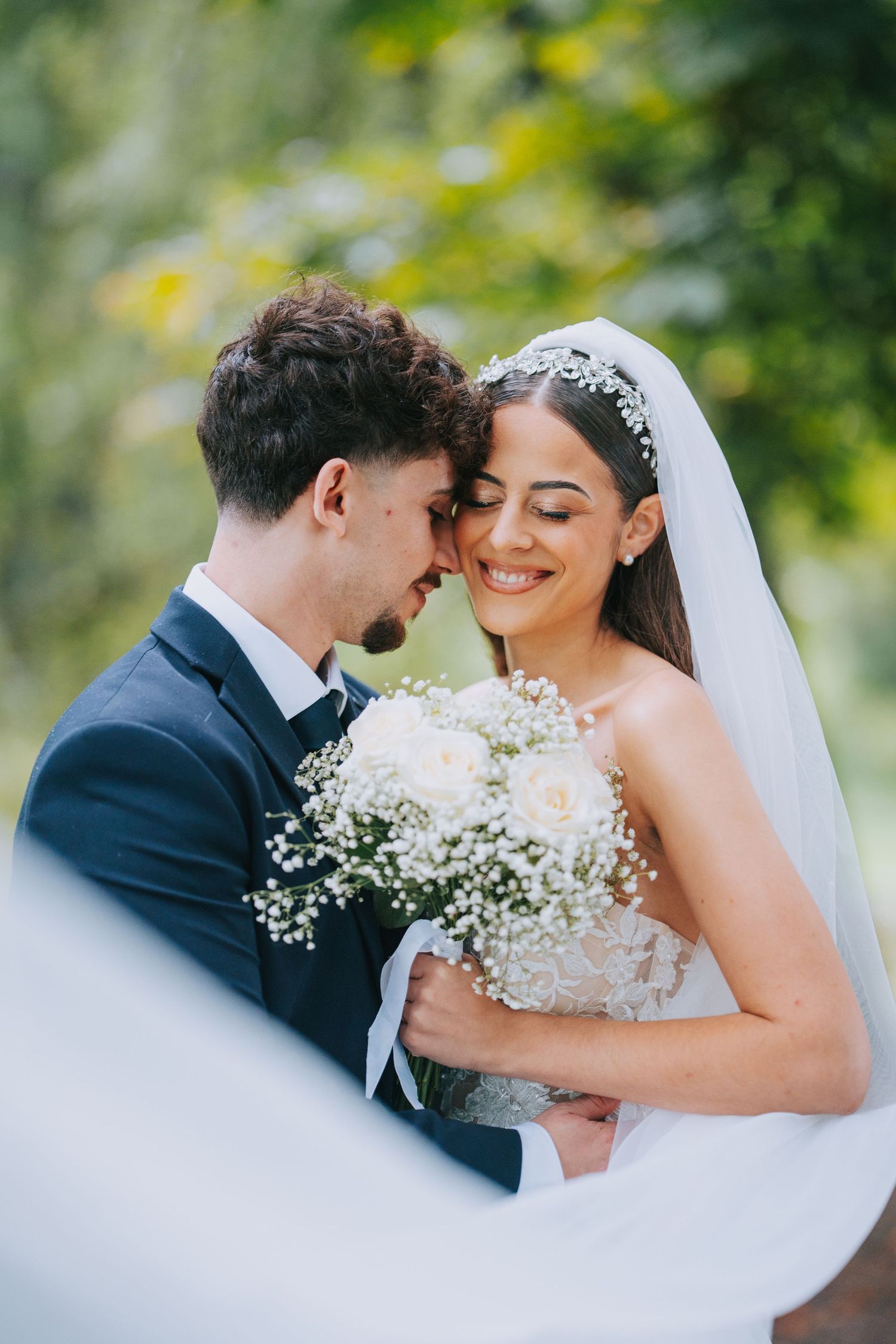 A joyful wedding couple embraces outdoors while holding a delicate bouquet of baby's breath flowers against a blurred green backdrop.