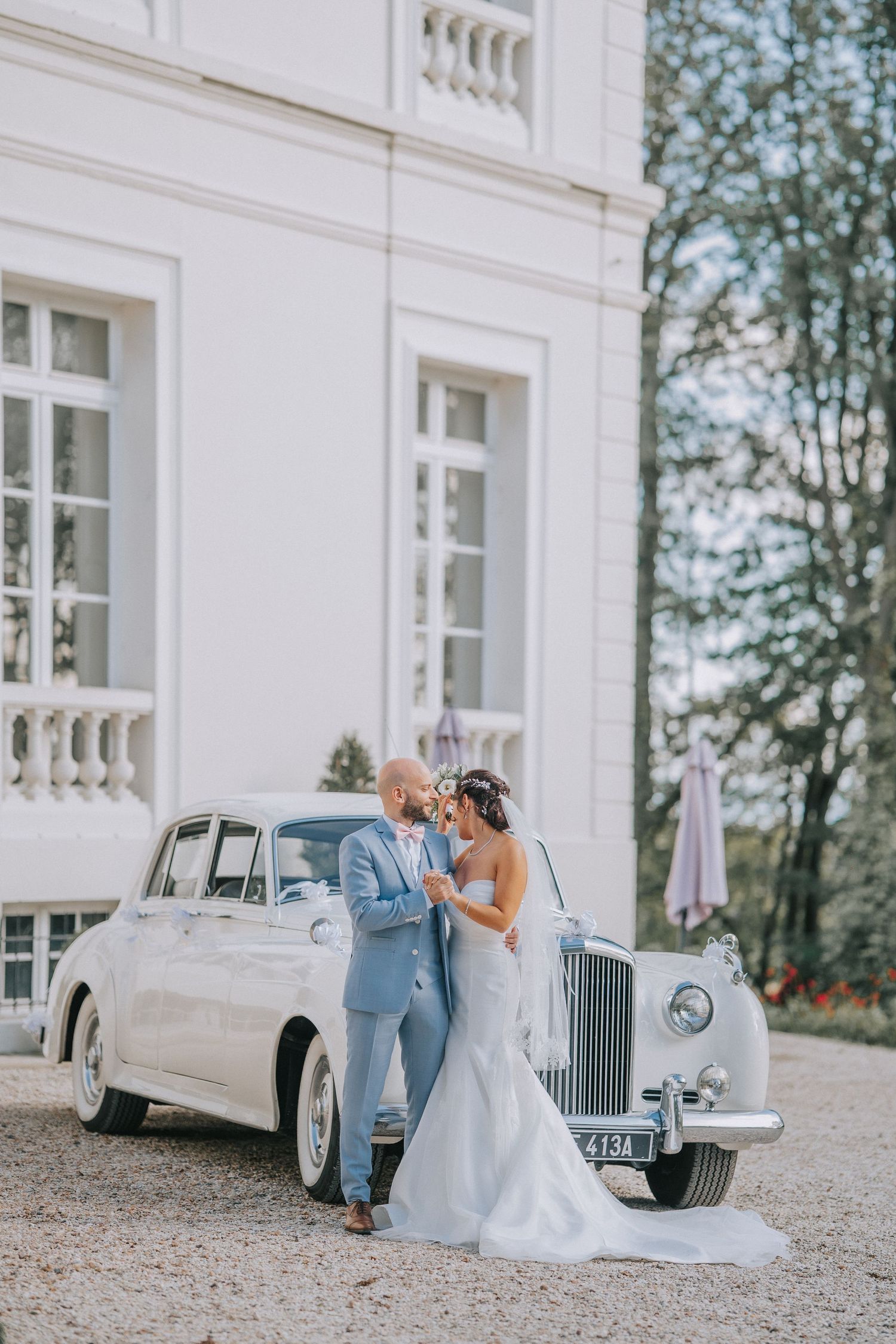 Newlyweds share romantic moment beside vintage white Rolls Royce at elegant mansion wedding venue.