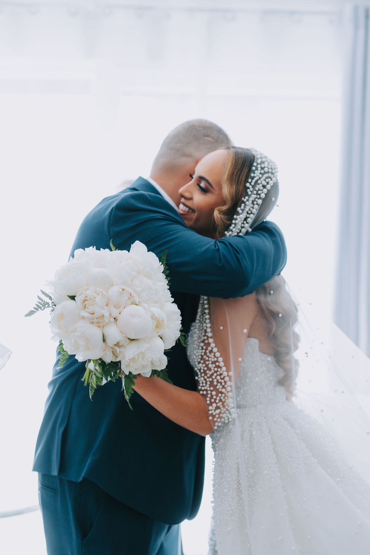 A tender embrace between a bride in a beaded wedding gown and someone in a navy suit while holding a white peony bouquet.