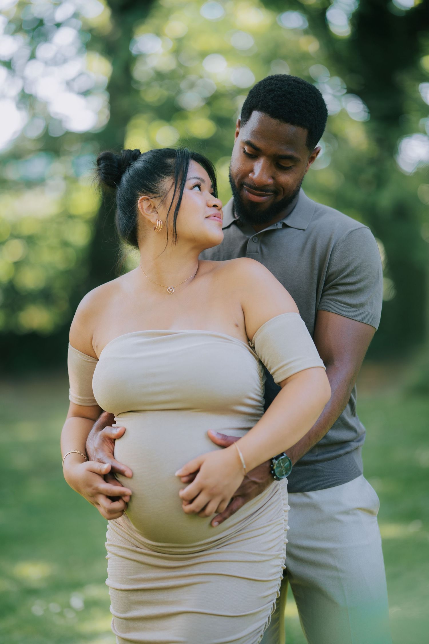A couple shares a tender moment outdoors during a maternity photoshoot in a sunny garden setting.