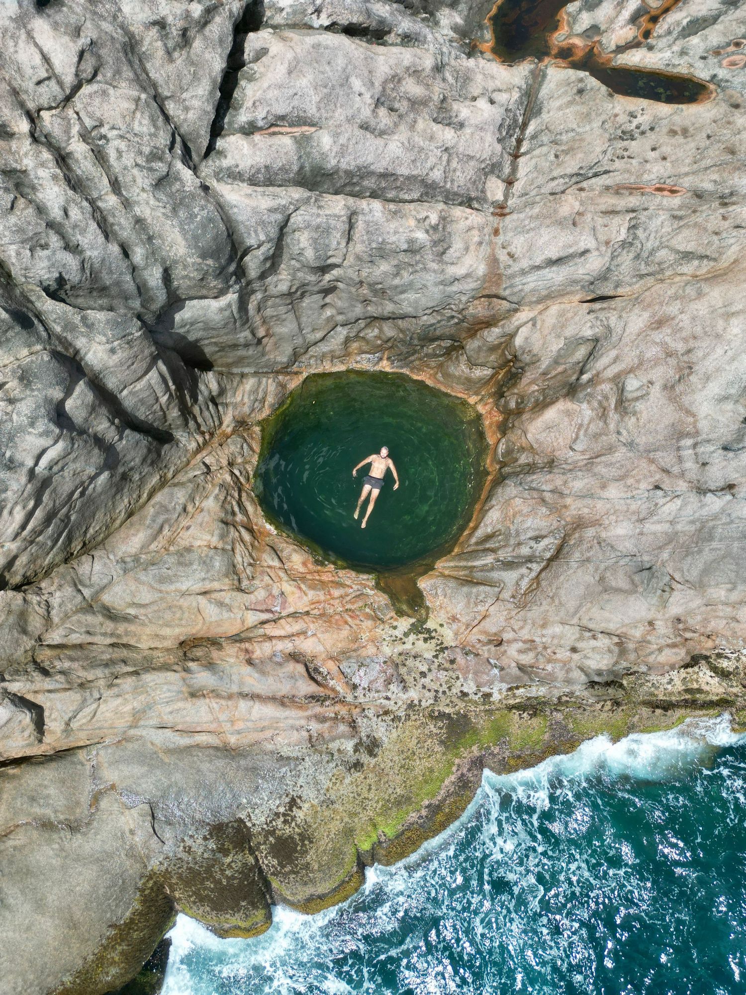 Aerial view of a natural rock pool with swimmer surrounded by rugged cliffs and crashing waves.