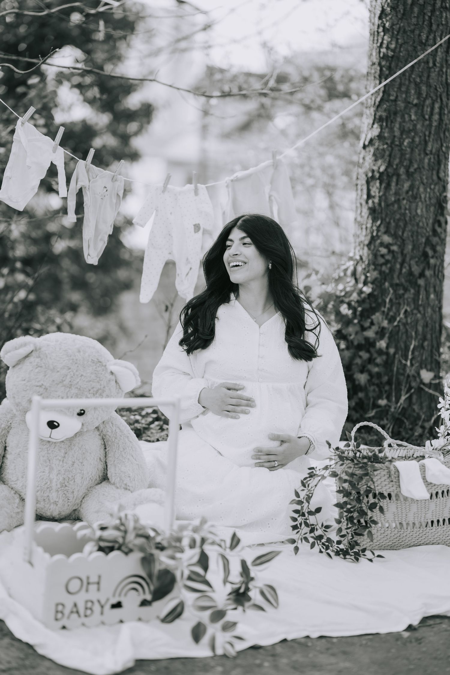 Black and white maternity photo with baby clothes on clothesline and teddy bear in outdoor setting.